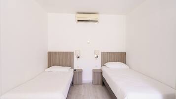Habitación de hotel con dos camas individuales, una mesita de noche pequeña con lámpara, un equipo de aire acondicionado en la pared y un enchufe.