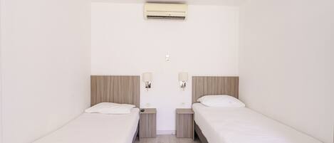Habitación de hotel con dos camas individuales, una mesita de noche pequeña con lámpara, un equipo de aire acondicionado en la pared y un enchufe.