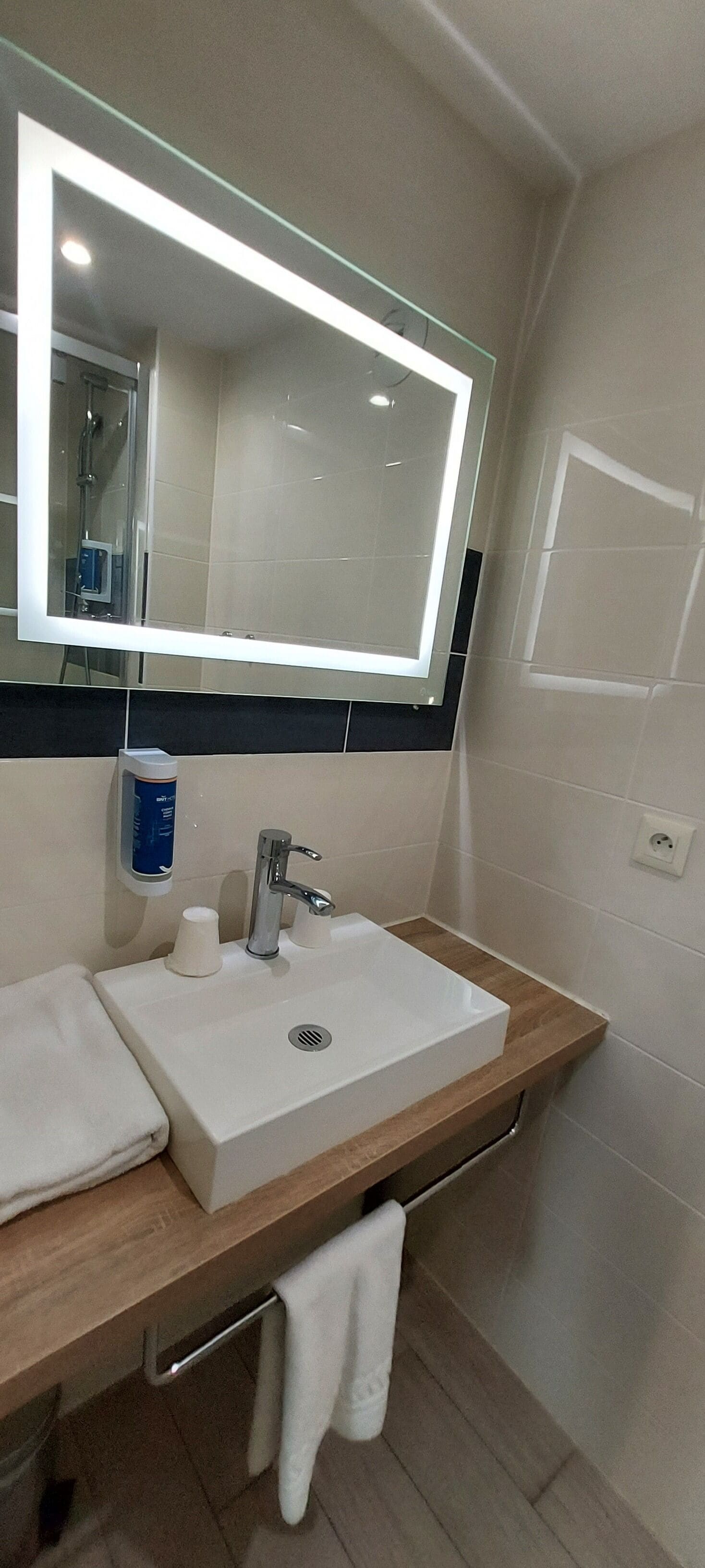 A modern bathroom with a white sink, a mirror with a light, a towel rack, and a soap dispenser.