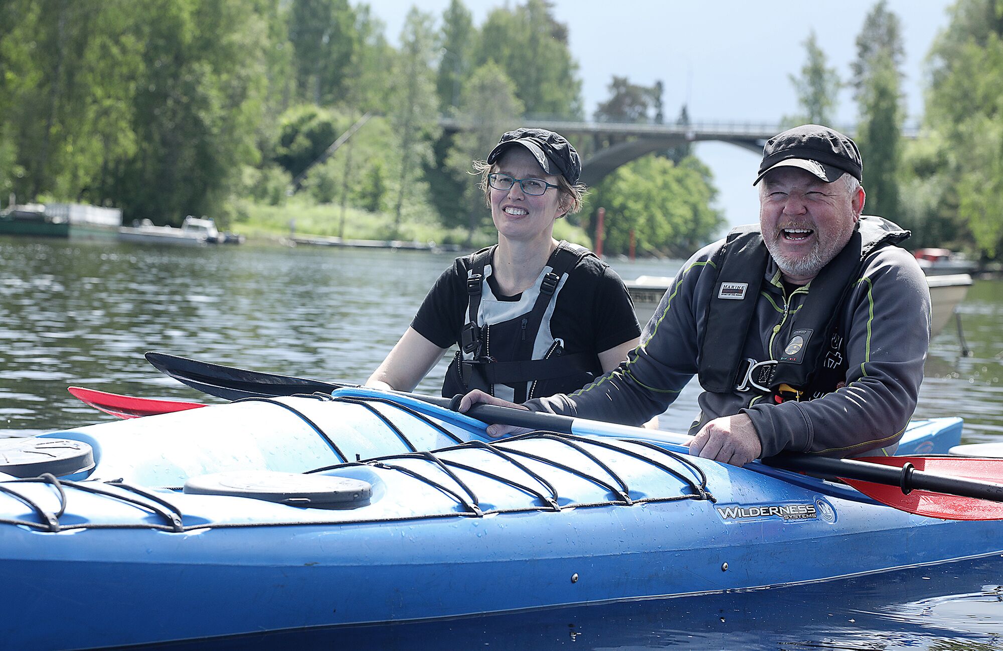 Two people on a blue kayak with a Wilderness Systems logo, a bridge, and trees in the background.