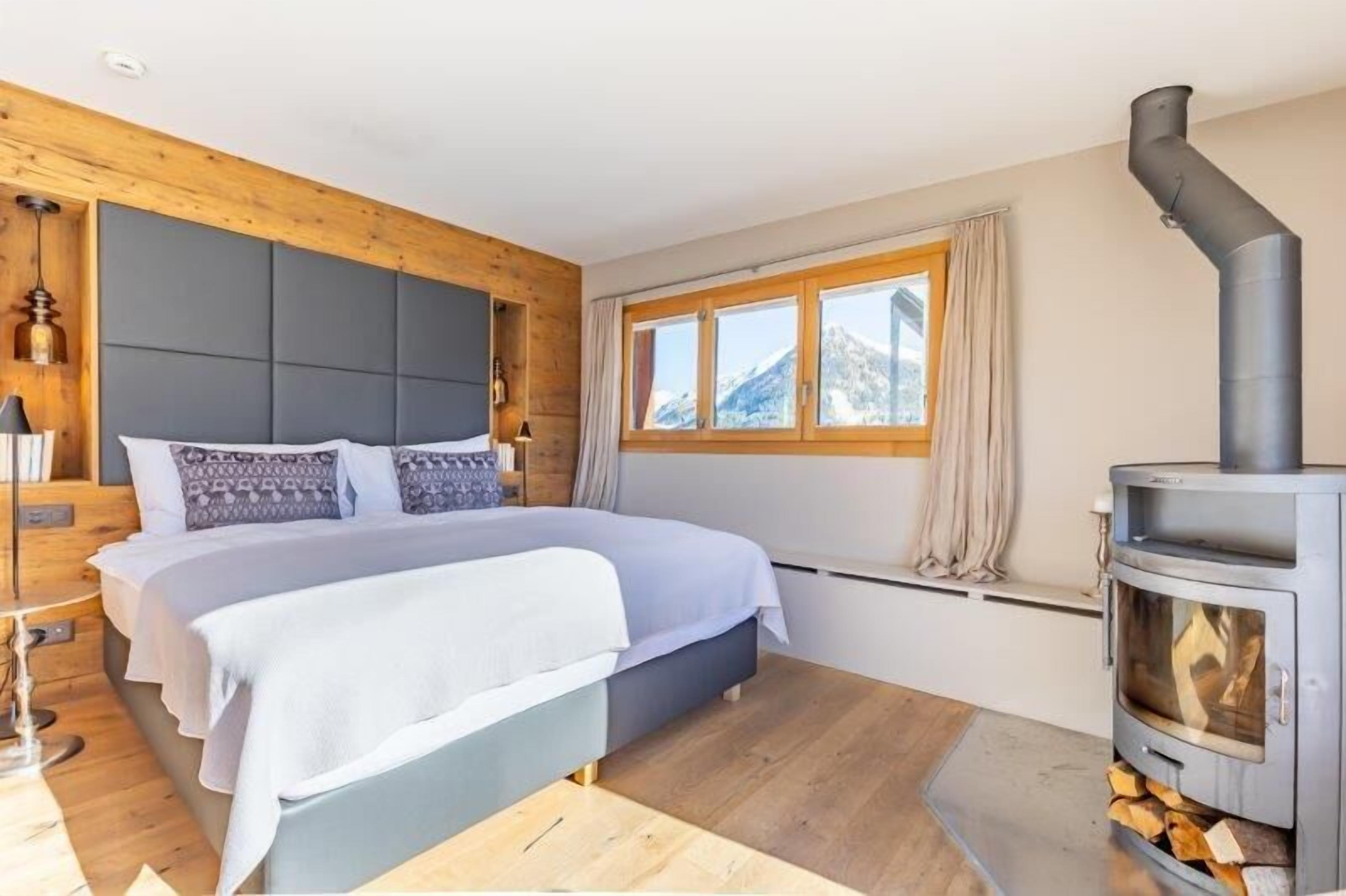 A bedroom with a bed, a wooden headboard, a window with curtains, and a wood-burning stove.