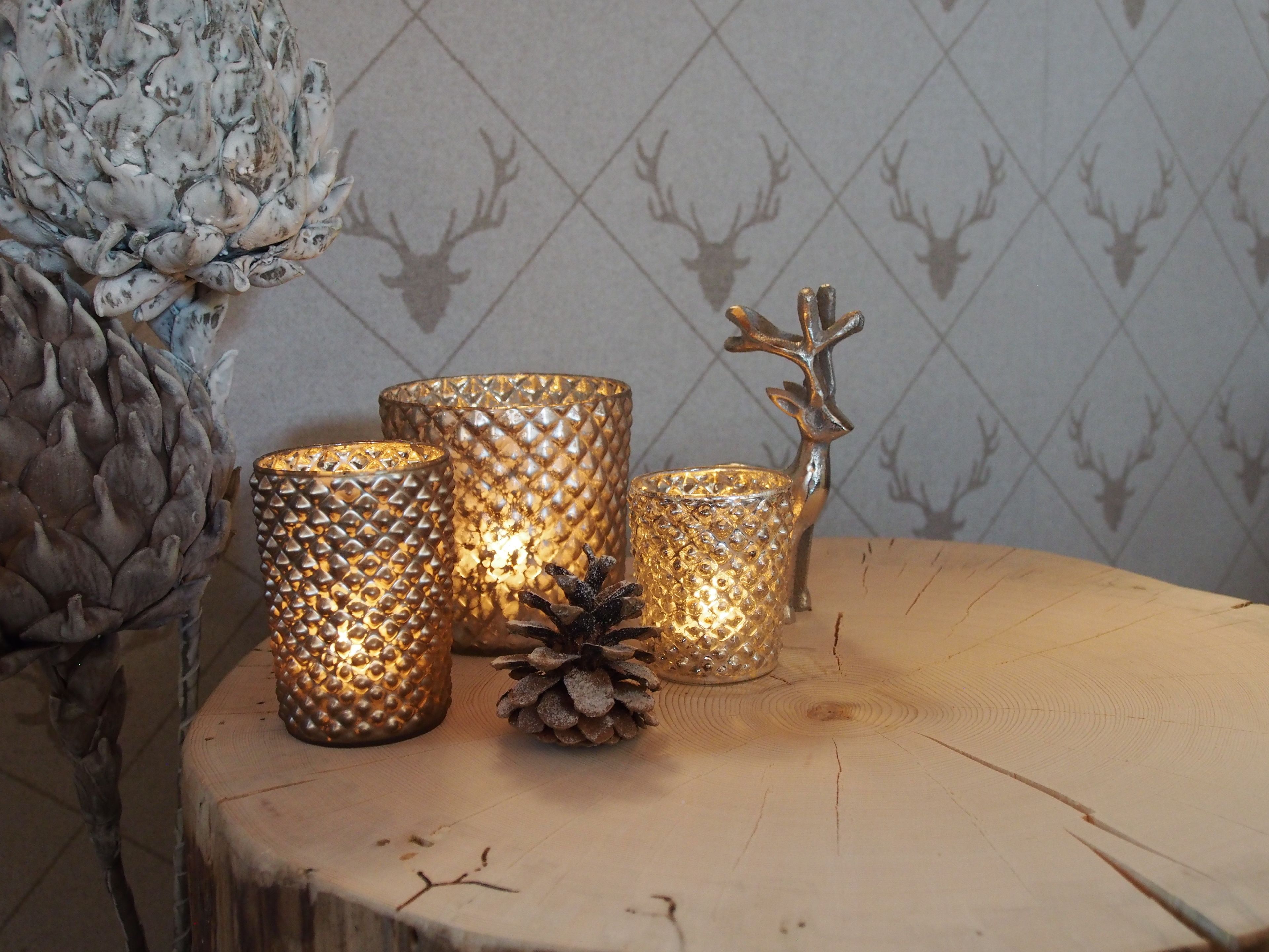 Three textured candle holders on a wooden surface with a pine cone and a deer head decoration.