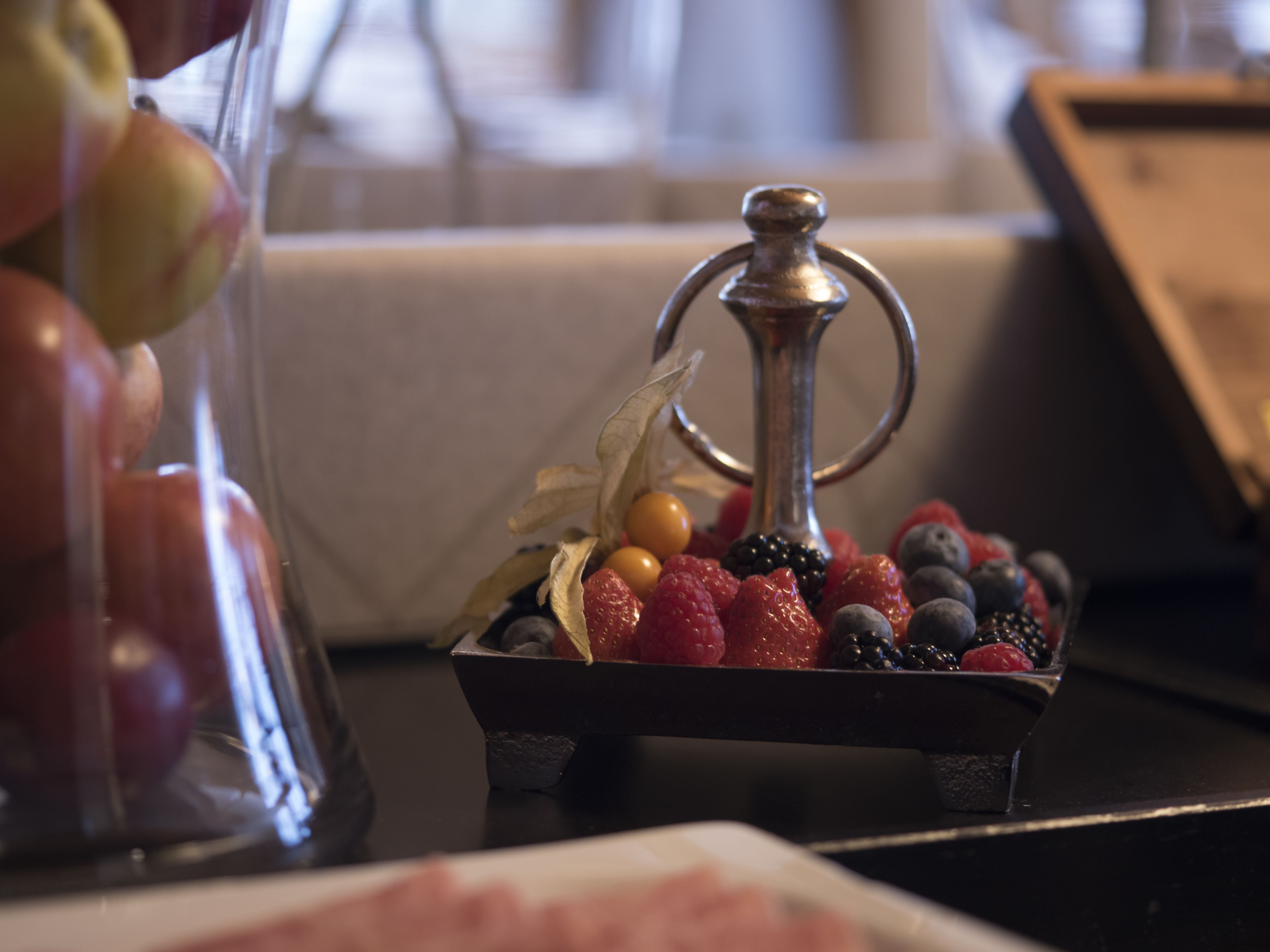 Una exhibición de frutas con un soporte metálico que sostiene diversas bayas y un recipiente de vidrio con manzanas.