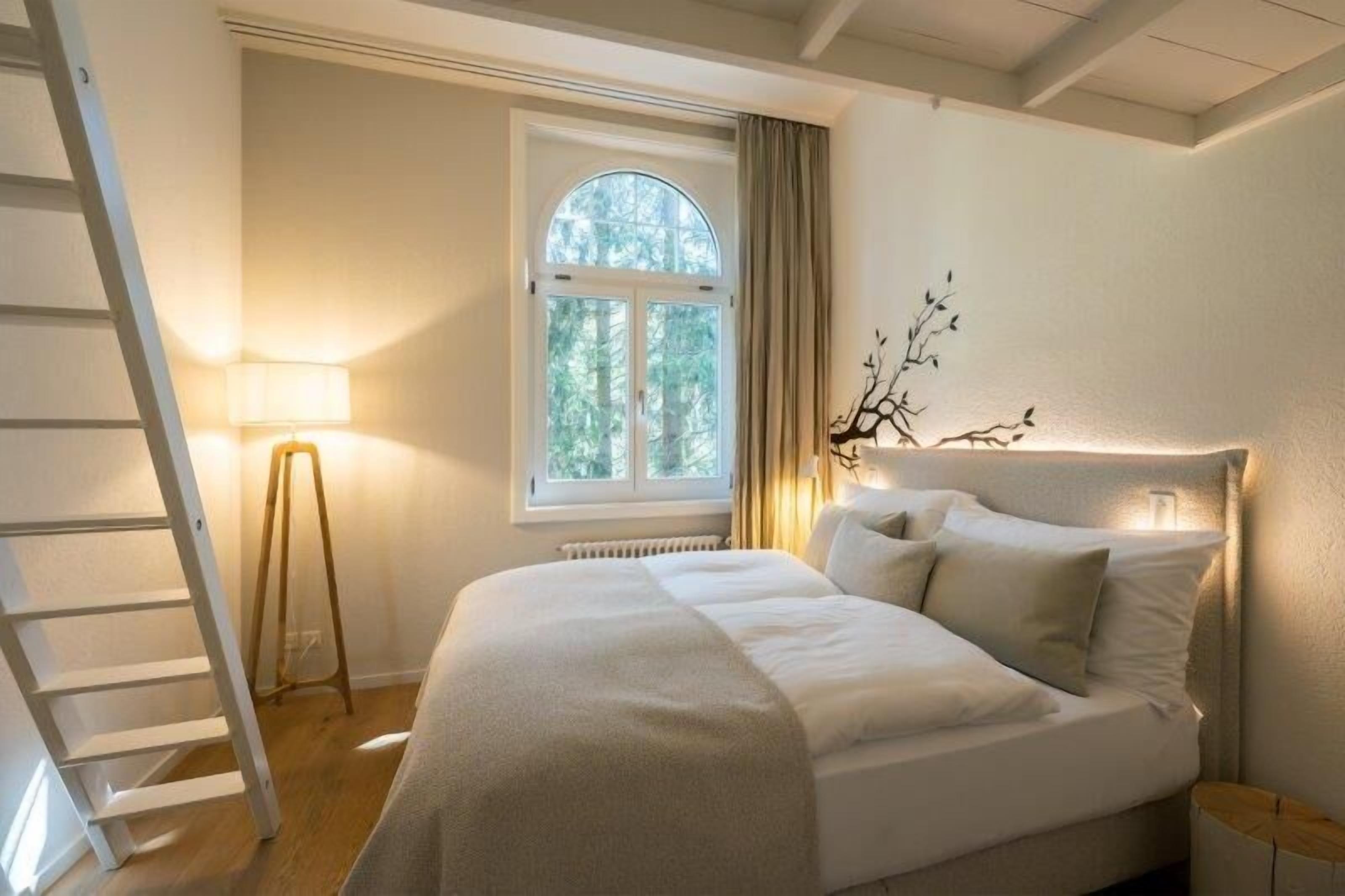 A bedroom with a bed, a window with curtains, a floor lamp, and a ladder.