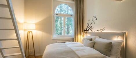 A bedroom with a bed, a window with curtains, a floor lamp, and a ladder.
