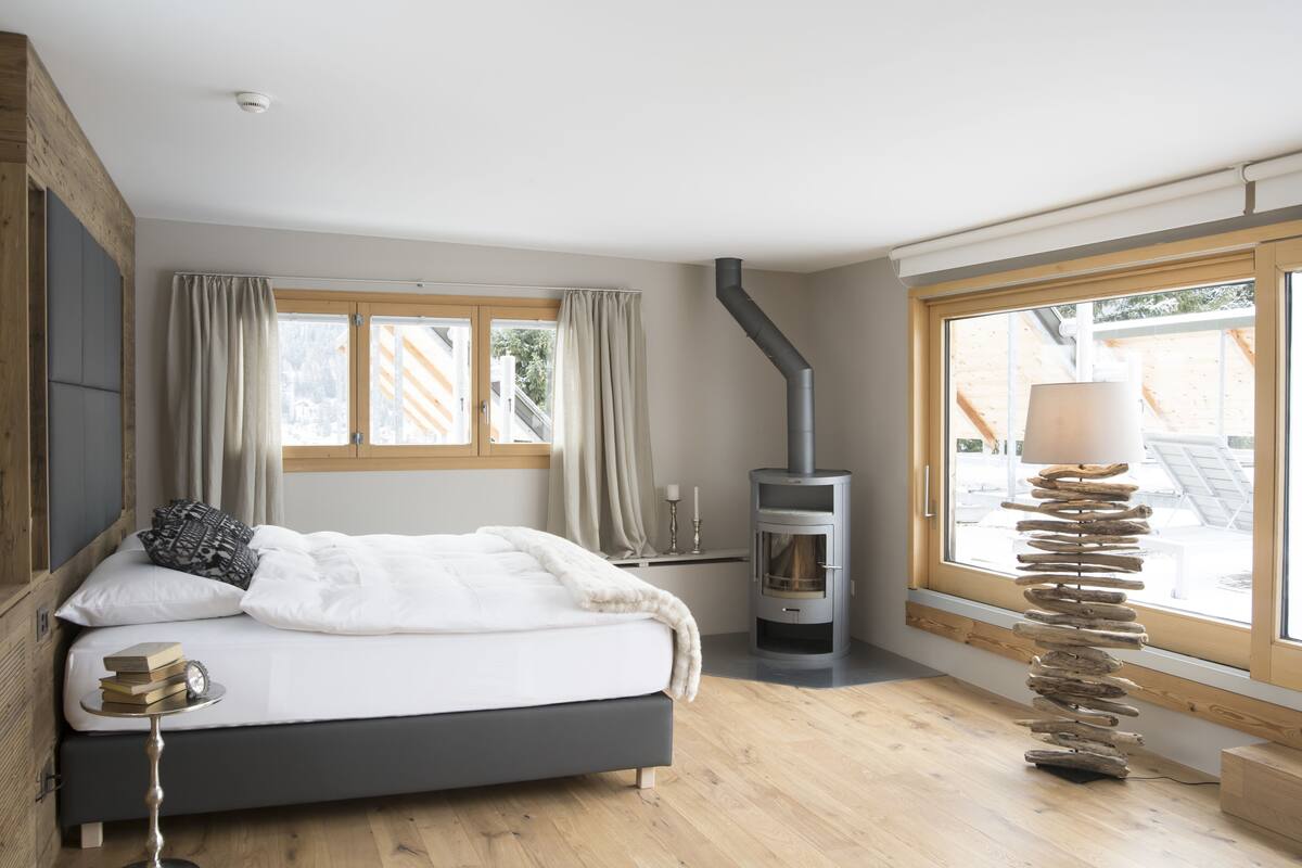 A bedroom with a bed, a wooden floor, a window with curtains, a fireplace, and a lamp.