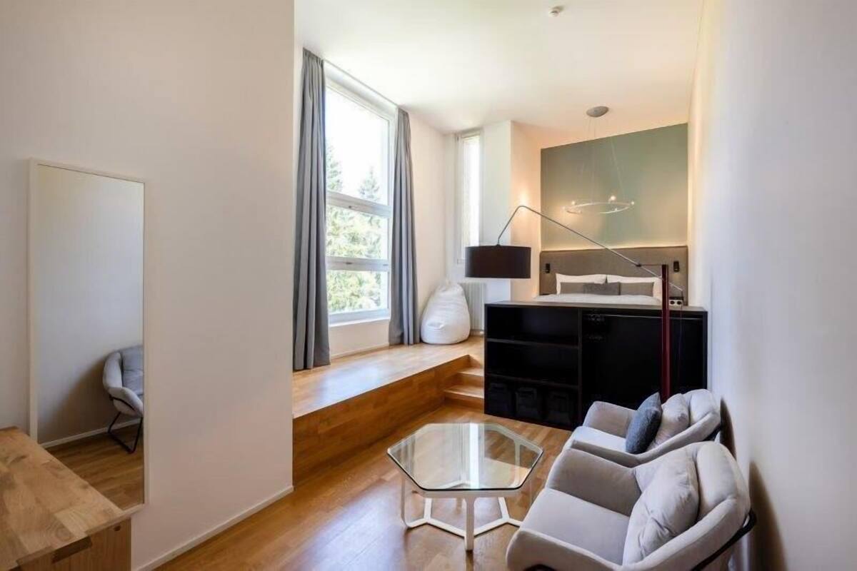 A modern living room with a glass coffee table, a sofa, a chair, and a dining area with a black cabinet and a lamp.