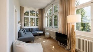 Un salon moderne avec un canapé d’angle gris, une table basse ronde, un téléviseur à écran plat, une lampe et de grandes fenêtres avec des rideaux.