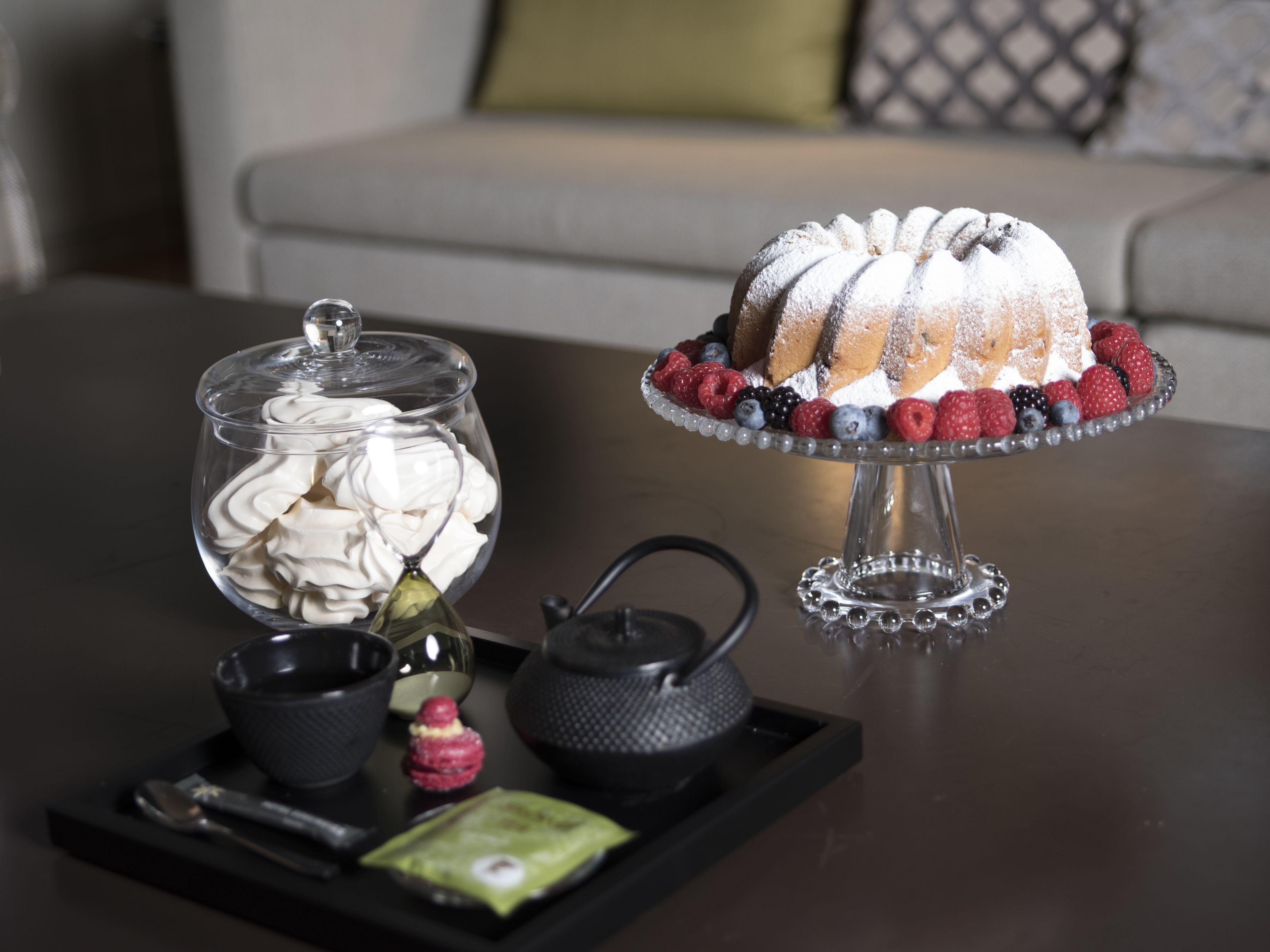 A dessert display with a bundt cake on a stand, a glass jar of whipped cream, and a tray with tea set and snacks.
