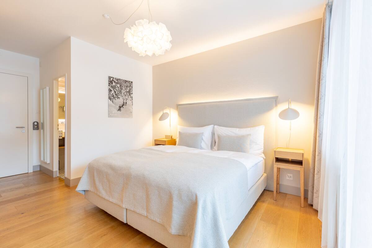 A modern bedroom with a large bed, bedside tables, a hanging light fixture, and a door leading to a bathroom.