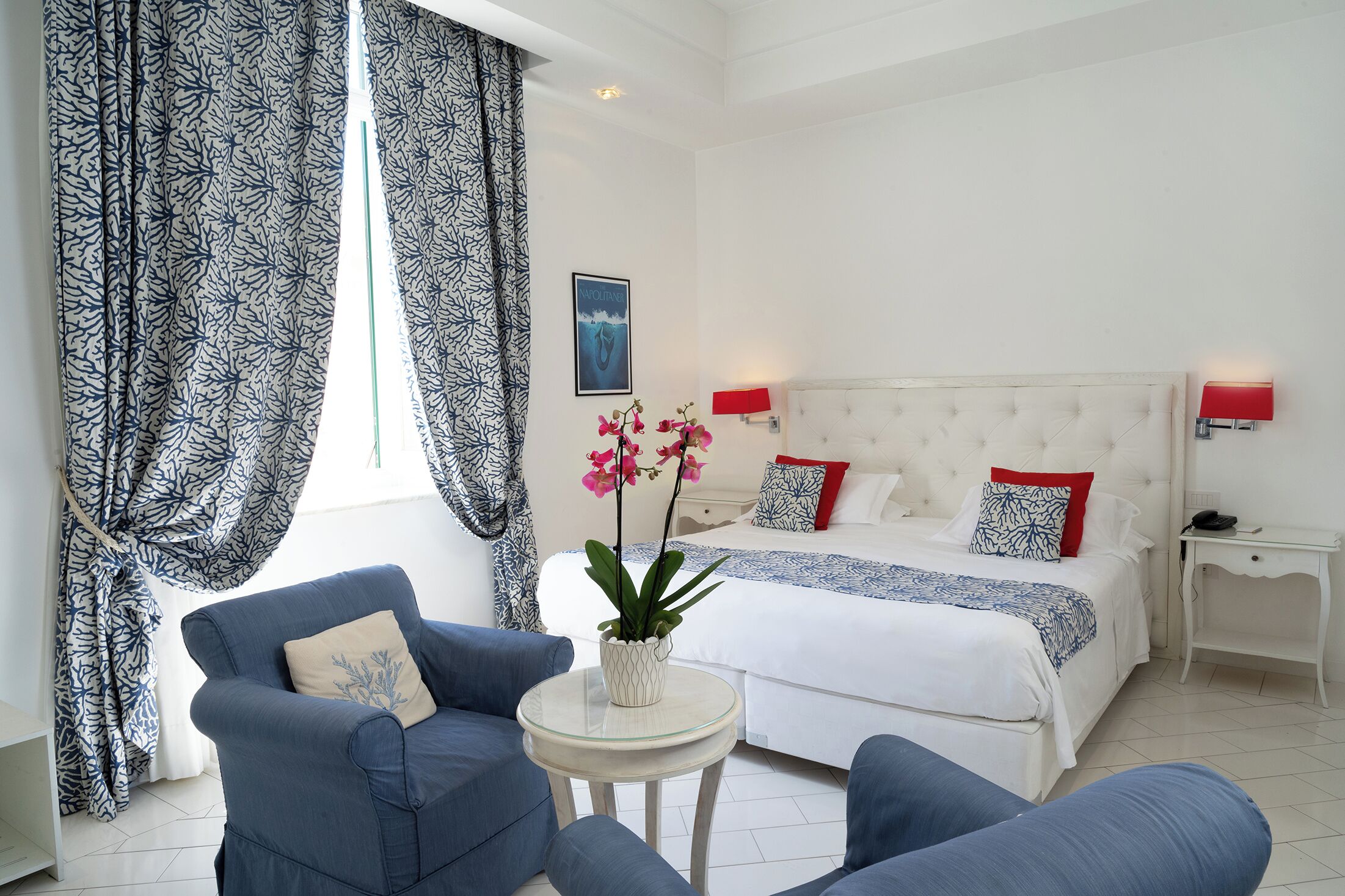 Ein Schlafzimmer mit einem Bett, zwei Nachttischen, einem Sessel, einem kleinen Tisch mit Pflanze und einem Fenster mit Vorhängen.