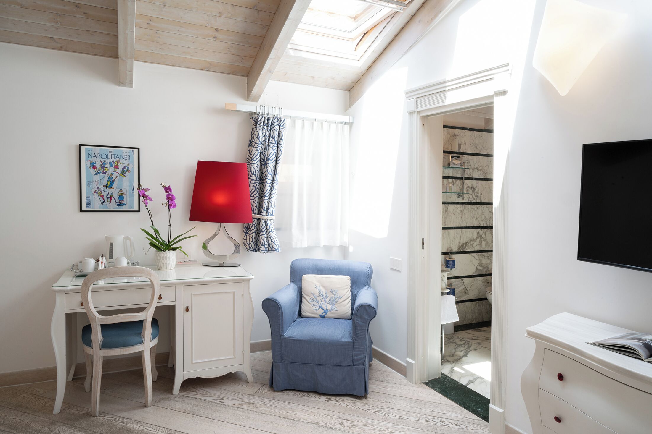 A room with a desk, chair, and armchair, a red lamp, a framed picture, and a TV on the wall.