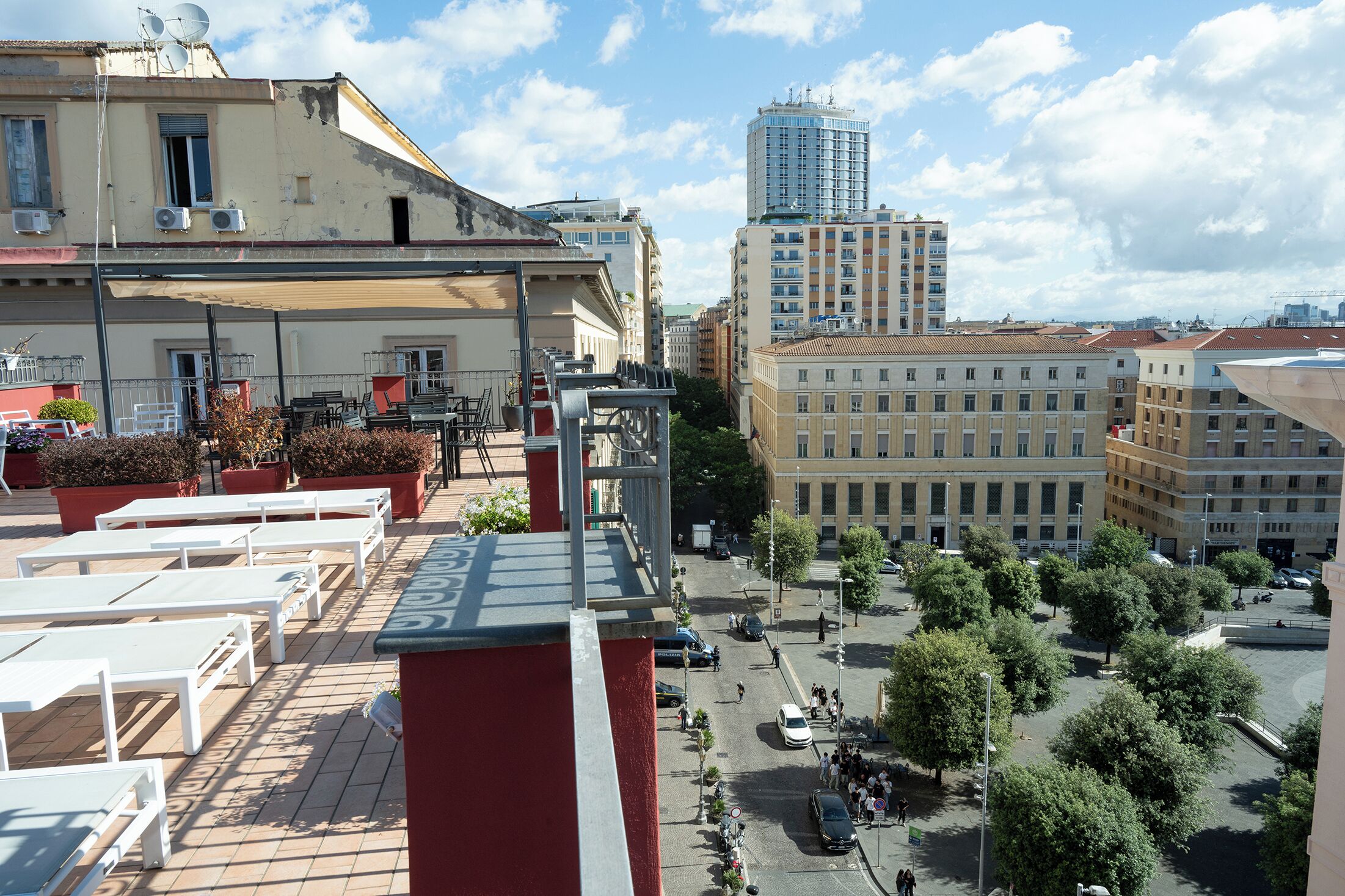 Terraza en la azotea con tumbonas, vista de un paisaje urbano que incluye un edificio alto, y una calle con árboles y peatones.