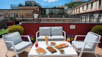 A rooftop terrace with outdoor seating, a table with food, and a view of buildings in the background.