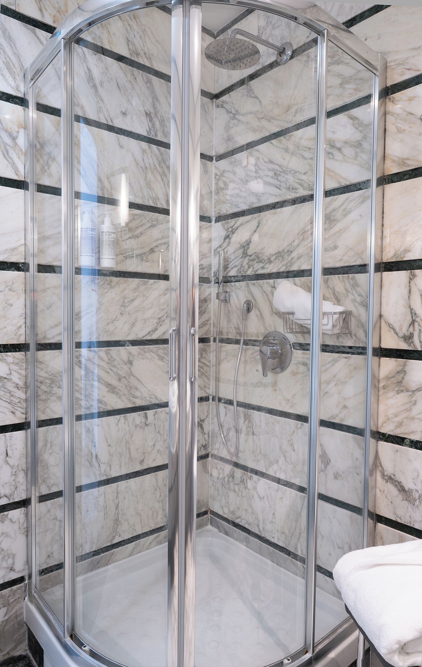 A glass-enclosed shower with a marble-tiled wall, a handheld showerhead, and a towel rack.