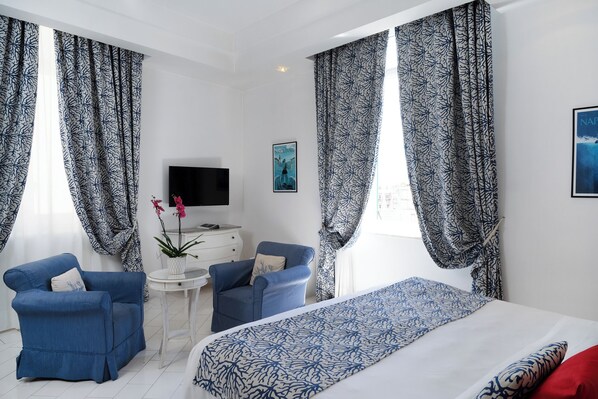 A hotel room with a bed, two blue armchairs, a small white table, a TV, and patterned curtains.