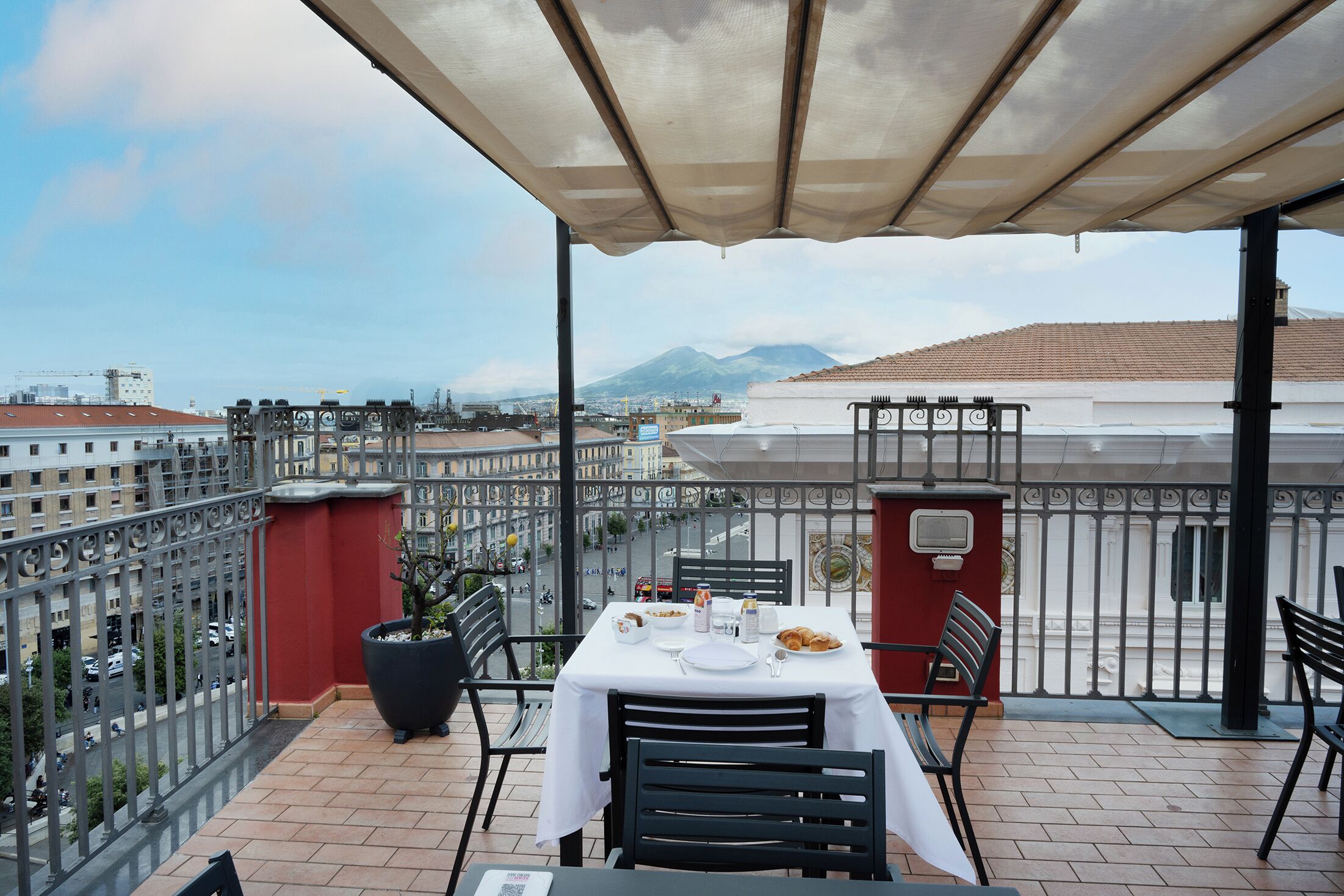 A balcony with a table set for dining, overlooking a cityscape with buildings and a mountain.