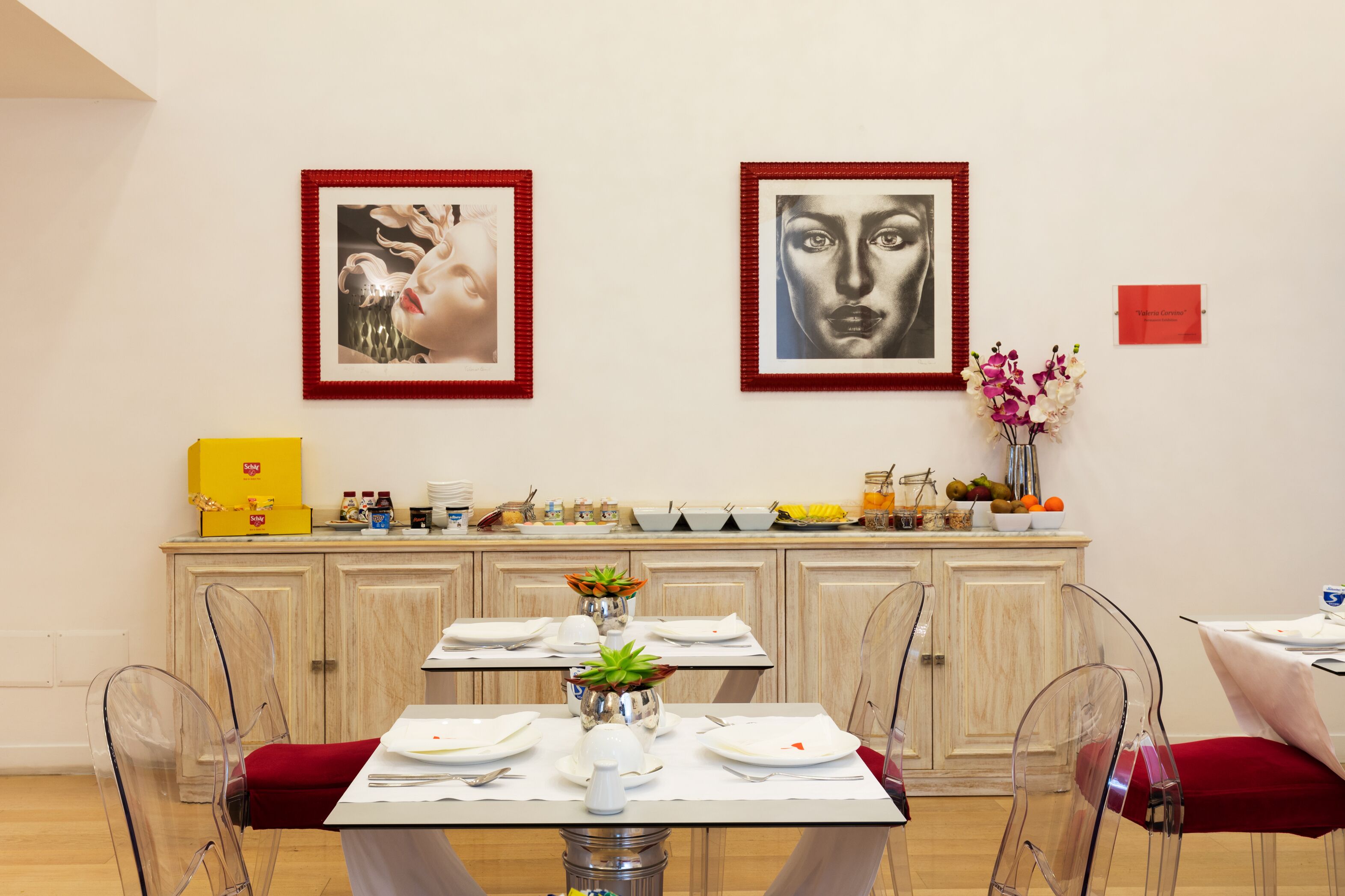 Un comedor con una mesa dispuesta para dos personas, sillas traslúcidas y un armario con utensilios de cocina.