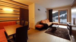 A modern living room with a wooden dining table, orange cushioned seating, a large window with a snowy view, and a dark rug. - Sissi Park (Oberwölz)