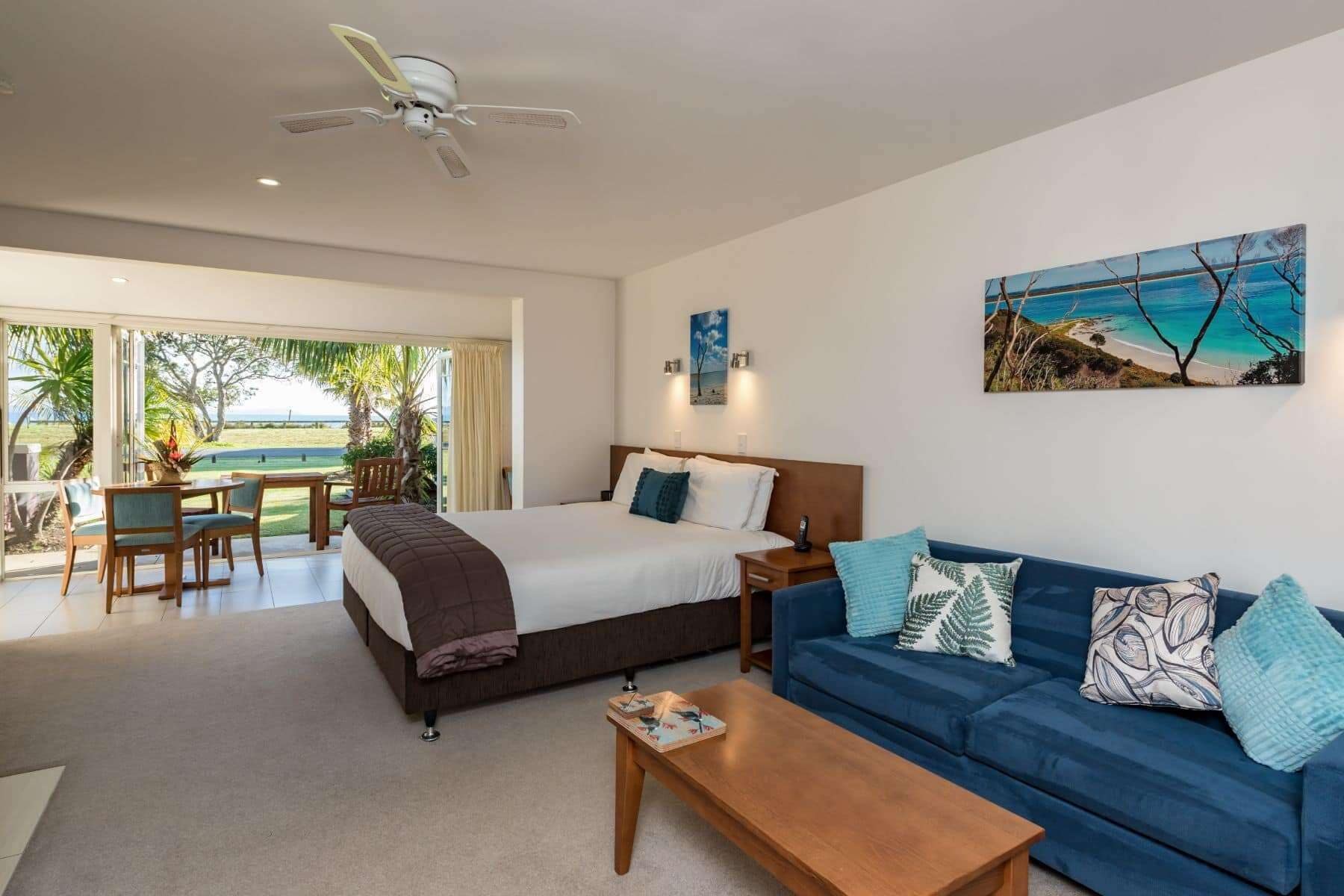 Quarto de hotel moderno com cama, sofá azul, mesa de centro e pintura de uma praia.