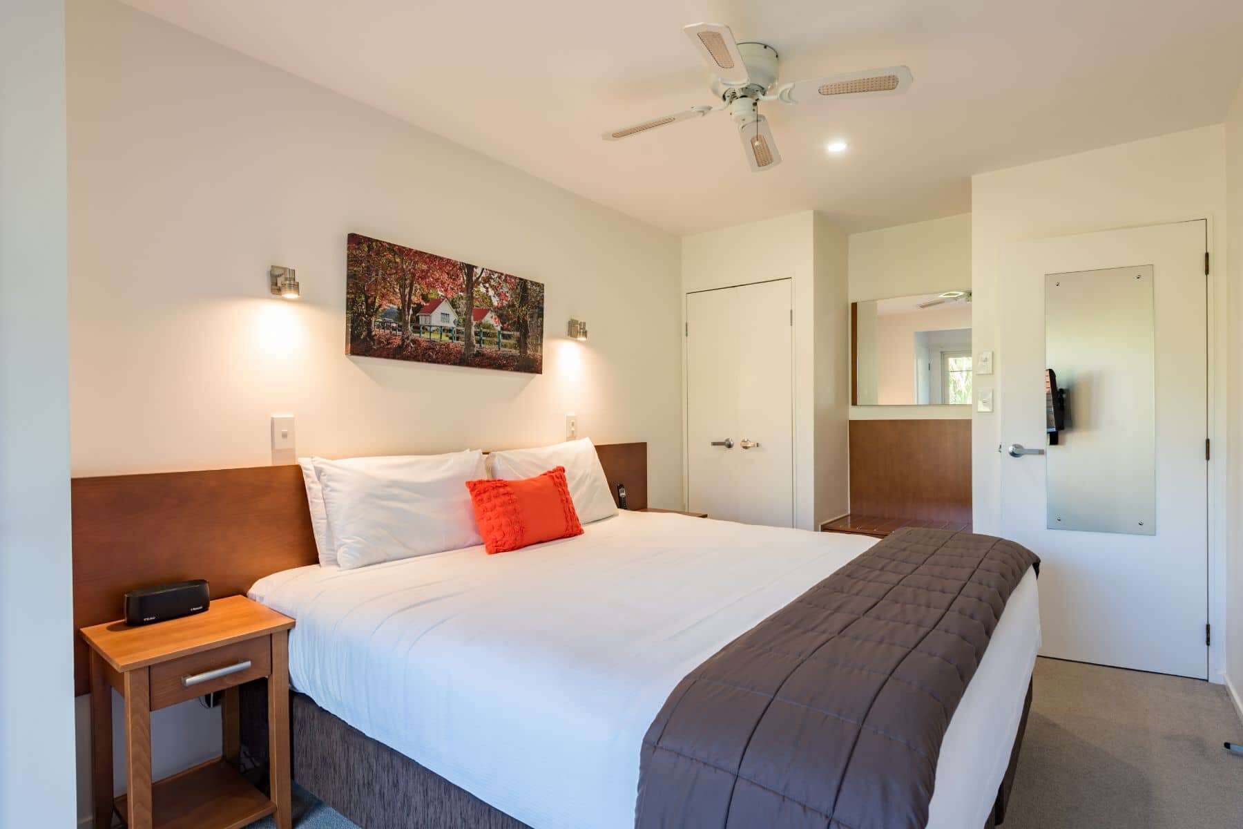 A hotel room with a large bed, a nightstand, a ceiling fan, a painting on the wall, and a door with a mirror.