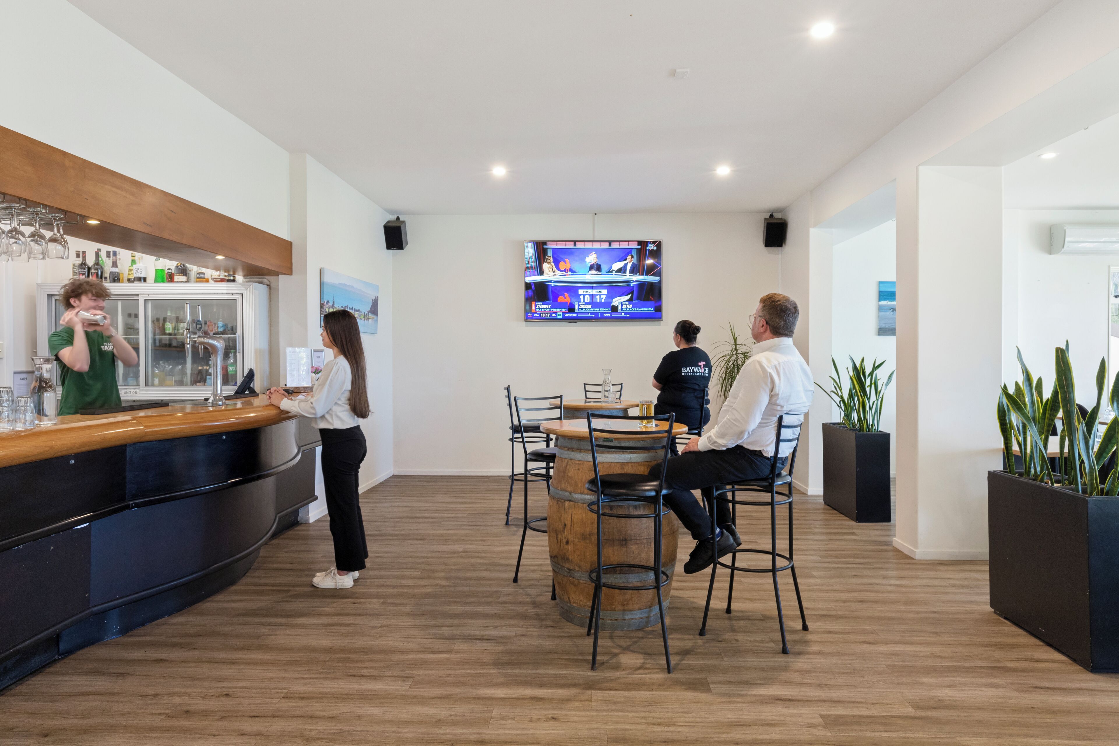 Zona de bar moderna con mostrador de madera, una pantalla mostrando deportes y un espacio de asientos con sillas de metal.
