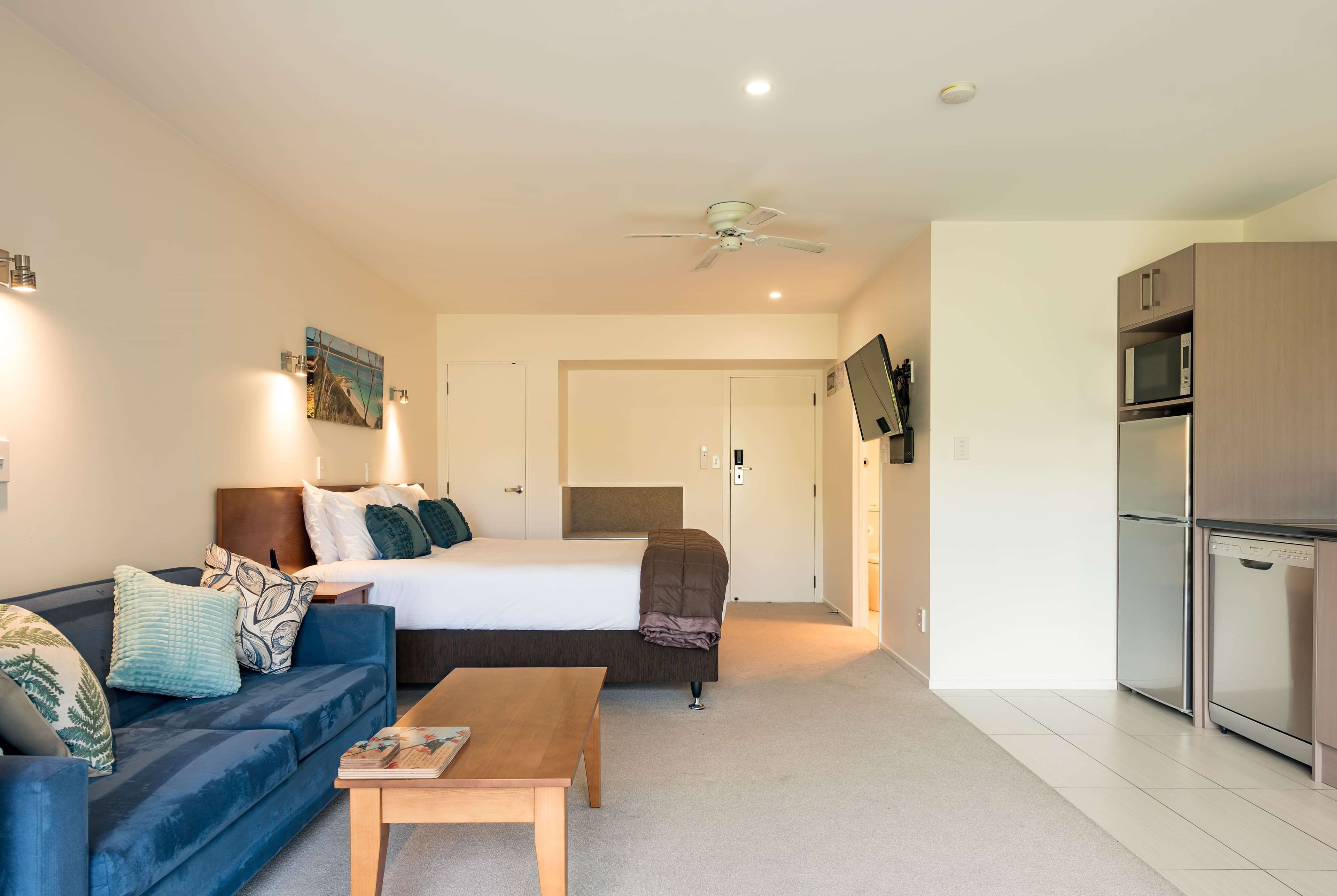 Ein Hotelzimmer mit einem Bett, einem blauen Sofa, einem Holztisch, einem Deckenventilator und einer Küchenzeile mit Mikrowelle und Kühlschrank.