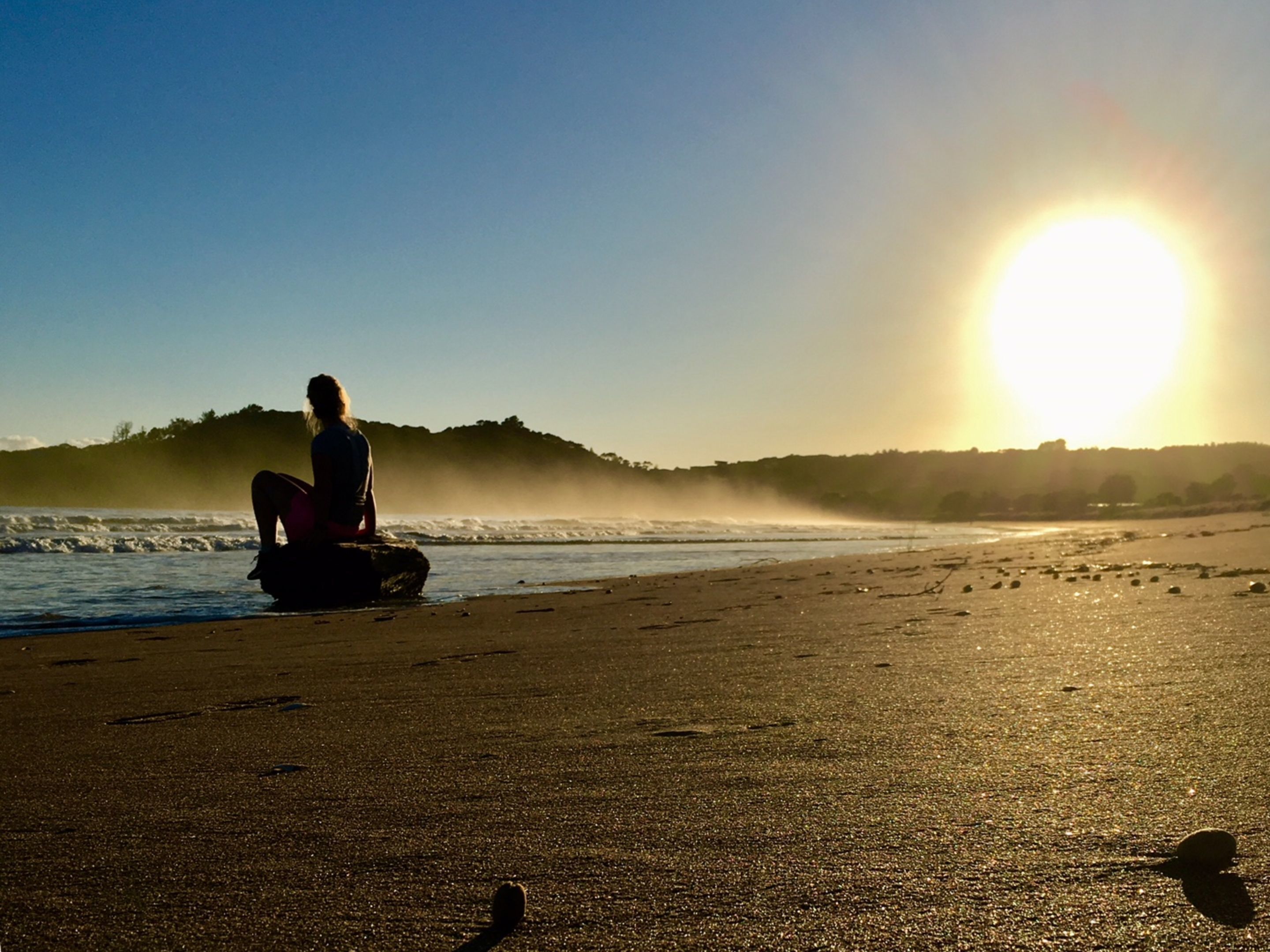 Eine Person sitzt an einem Felsen am Meer bei Sonnenuntergang.