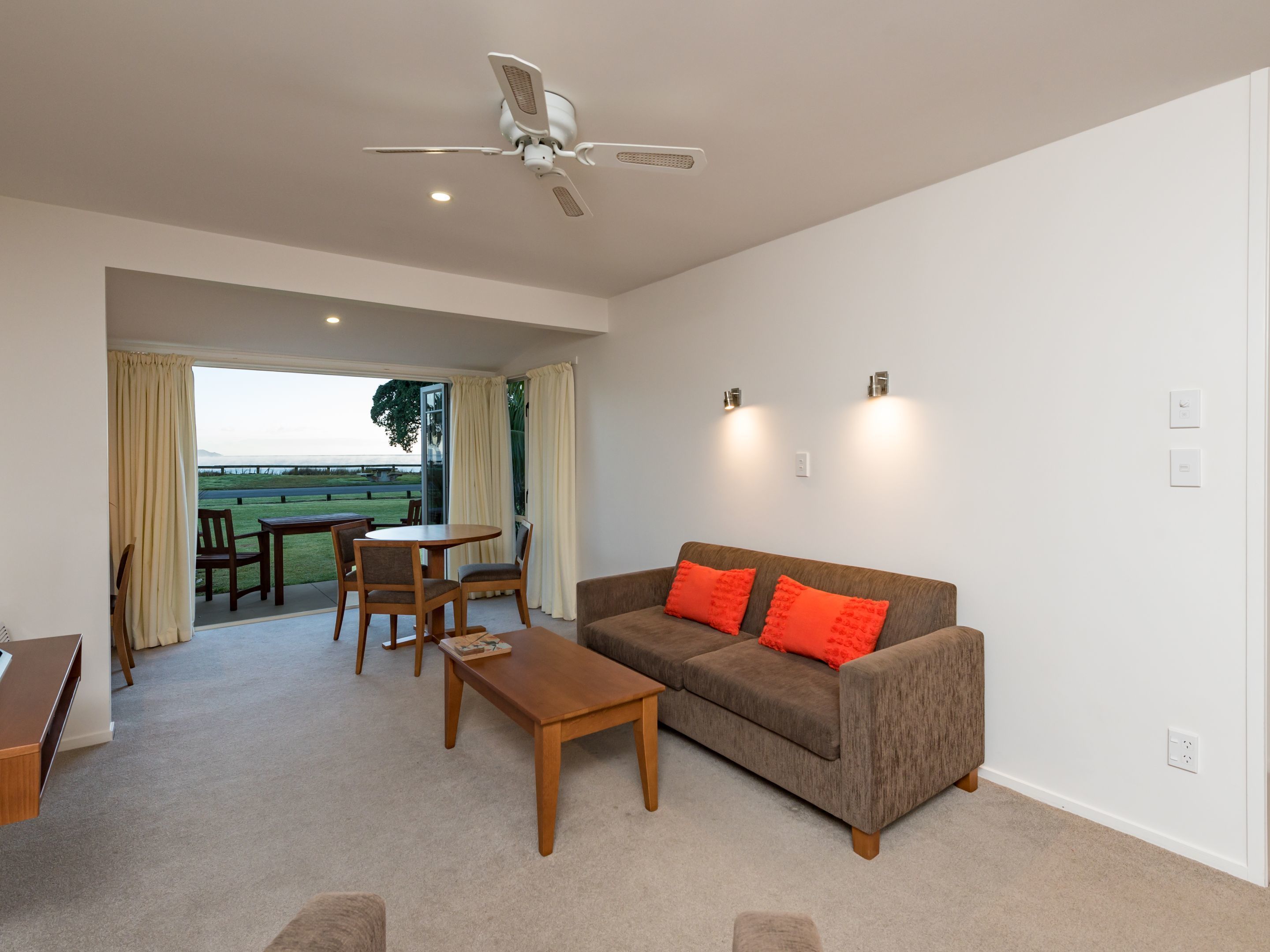 A spacious living room with a sofa, coffee table, and chairs, a ceiling fan, and a view of a grassy area through a sliding glass door.