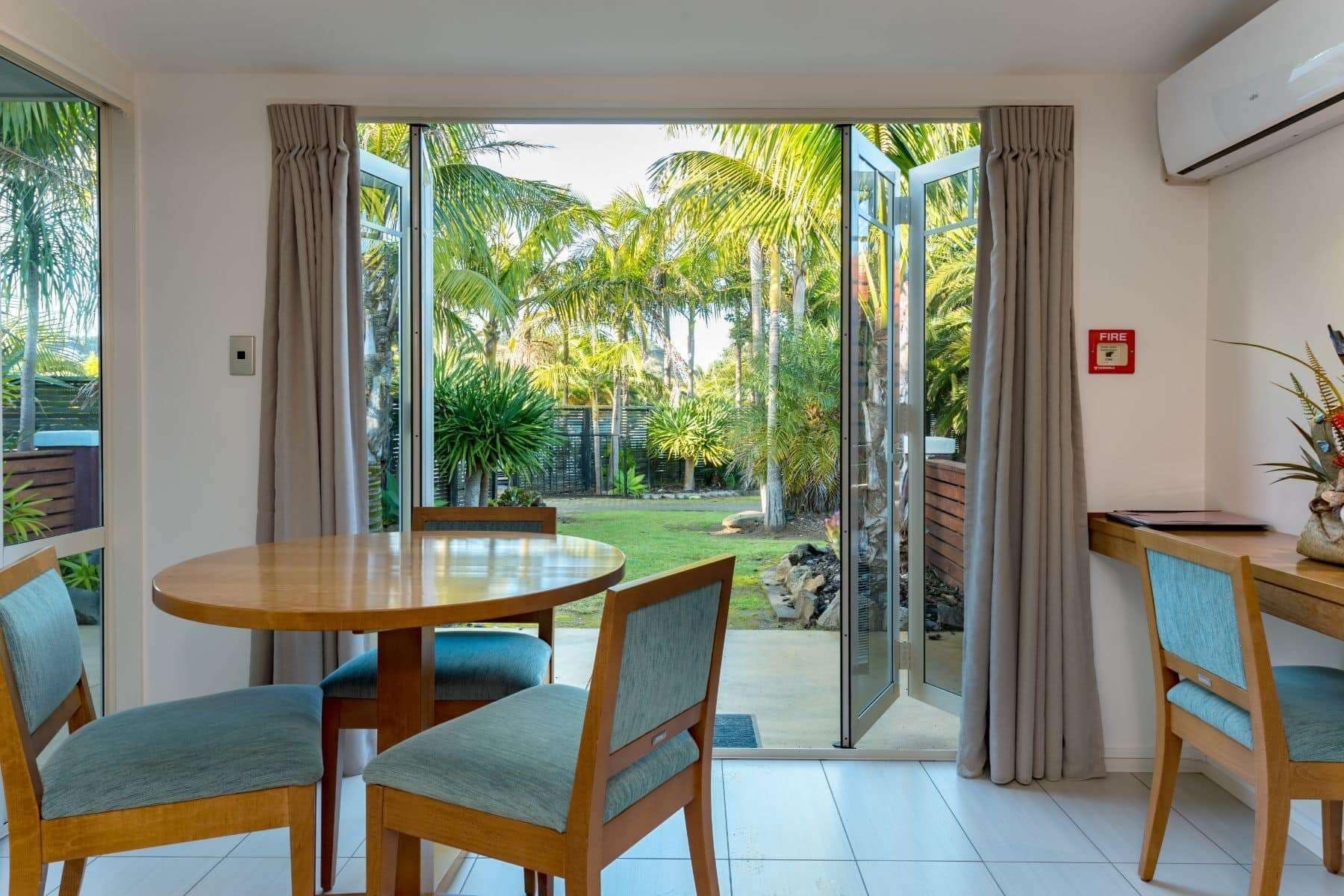 A room with a round wooden table and chairs, a fire safety sign, and a view of a garden with palm trees.