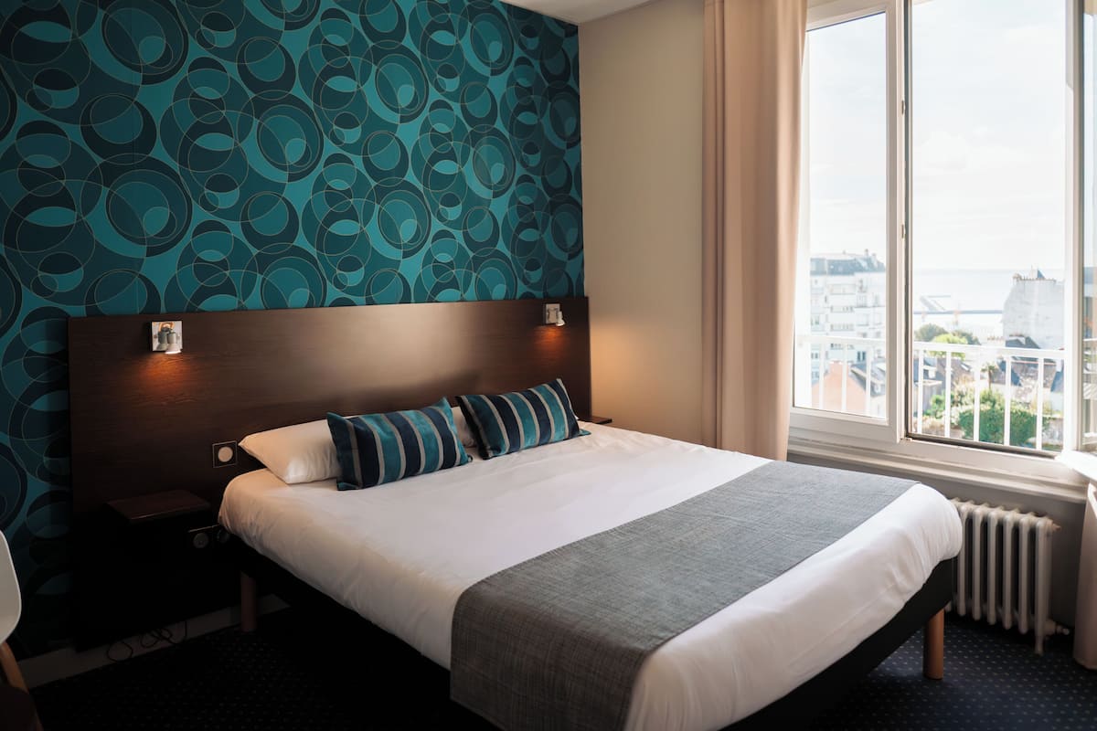 A hotel room with a large bed, a headboard with lights, a view of the city through a window, and a patterned wall.