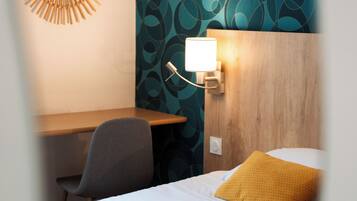 A hotel room with a bed, a desk, and a chair. There is a wall-mounted light fixture above the desk.