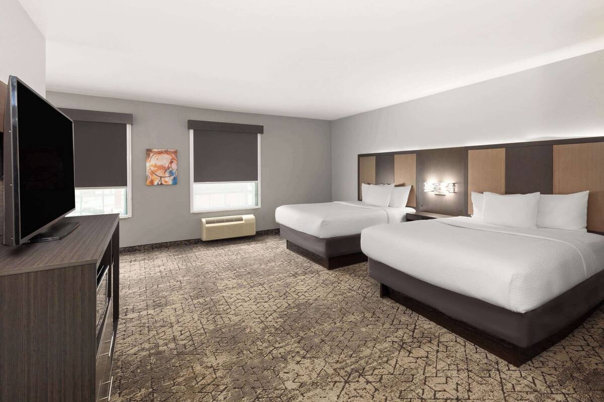 Une chambre d’hôtel avec deux lits, une télévision, un tableau et une fenêtre avec des stores.