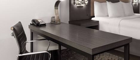 A hotel room with a desk, chair, bedside table, lamp, and a bed with pillows.