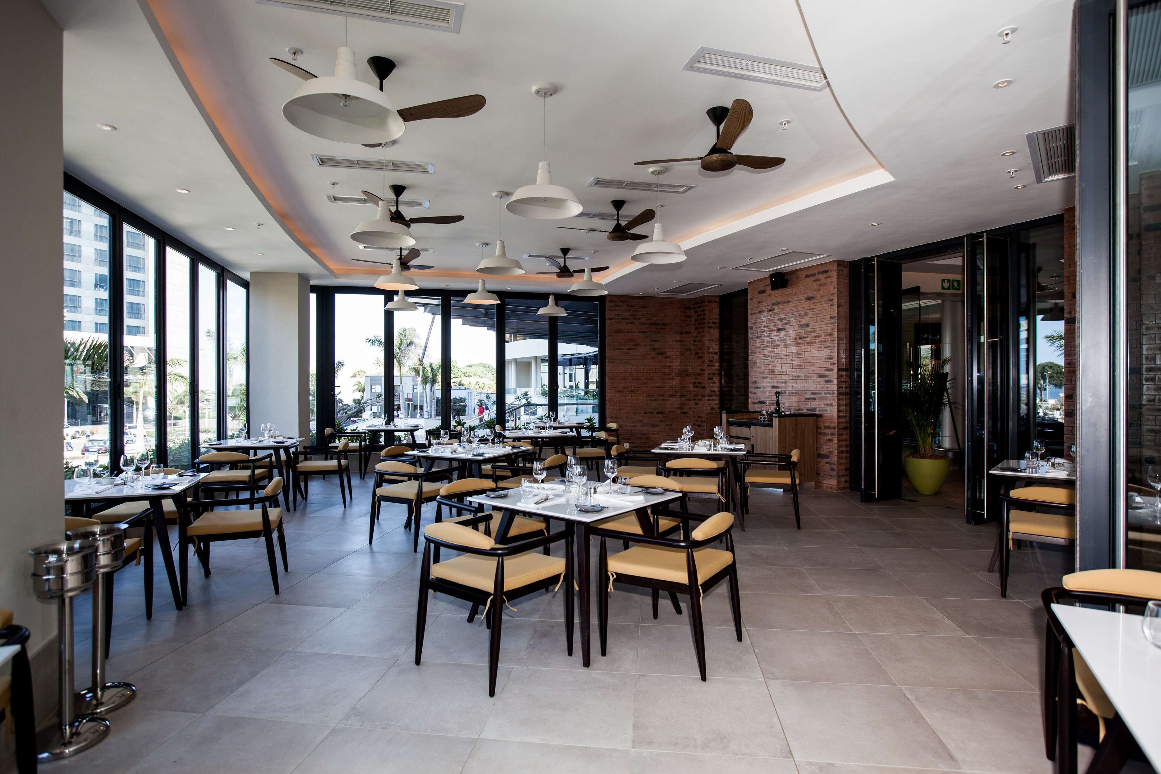 A spacious restaurant with multiple tables and chairs, ceiling fans, and large windows offering views of the outside.