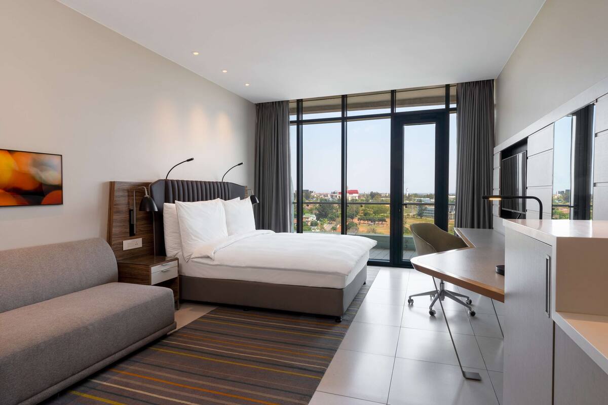 A modern hotel room with a bed, a desk, a sofa, and a large window offering a city view.