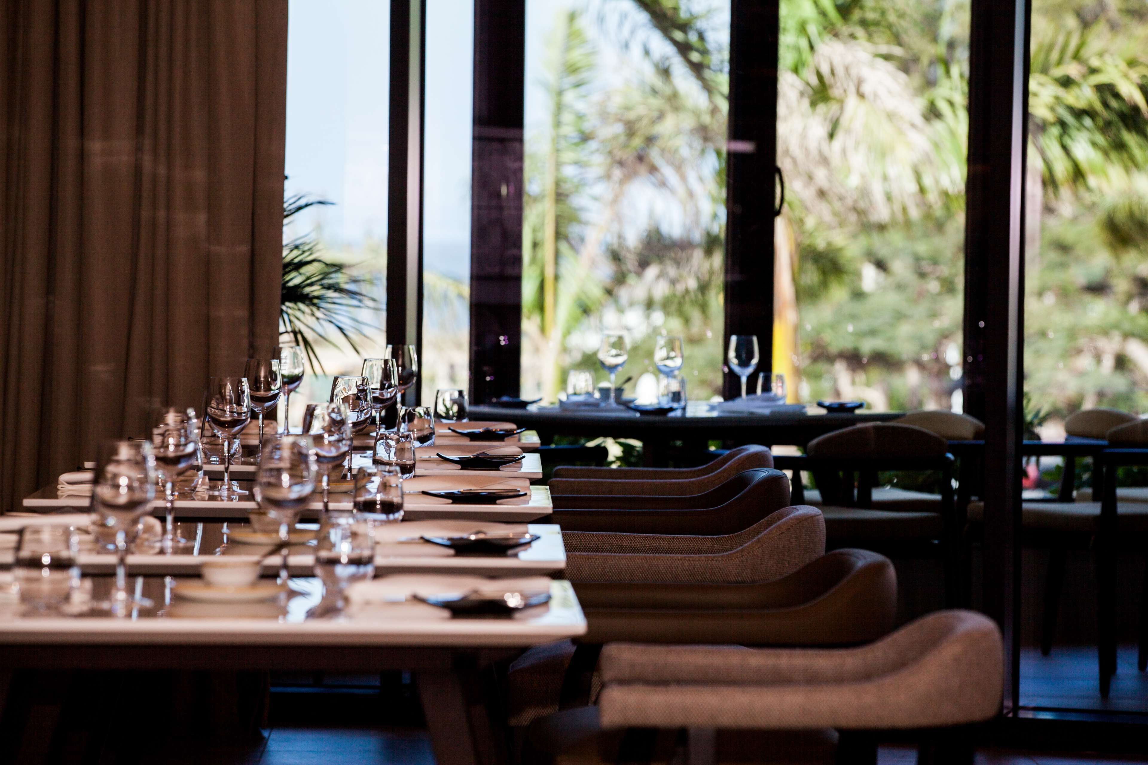 A restaurant with a long table set for dining, large windows, and a view of palm trees.