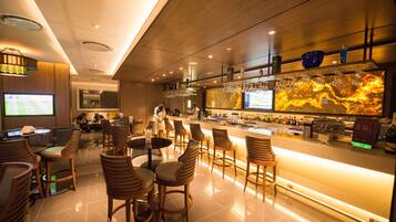 A modern bar area with wooden chairs and tables, a large TV screen, and a bar counter with various bottles and glasses.