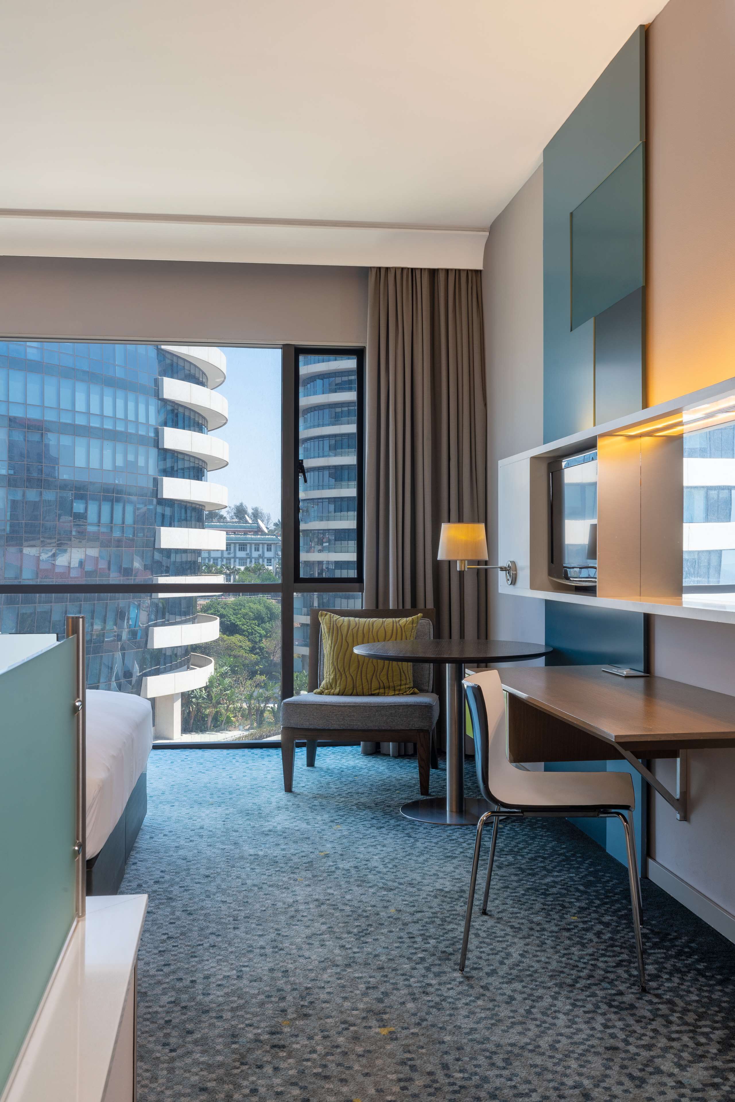 Habitación de hotel con cama, escritorio, silla, televisión y ventana grande con vistas a la ciudad.