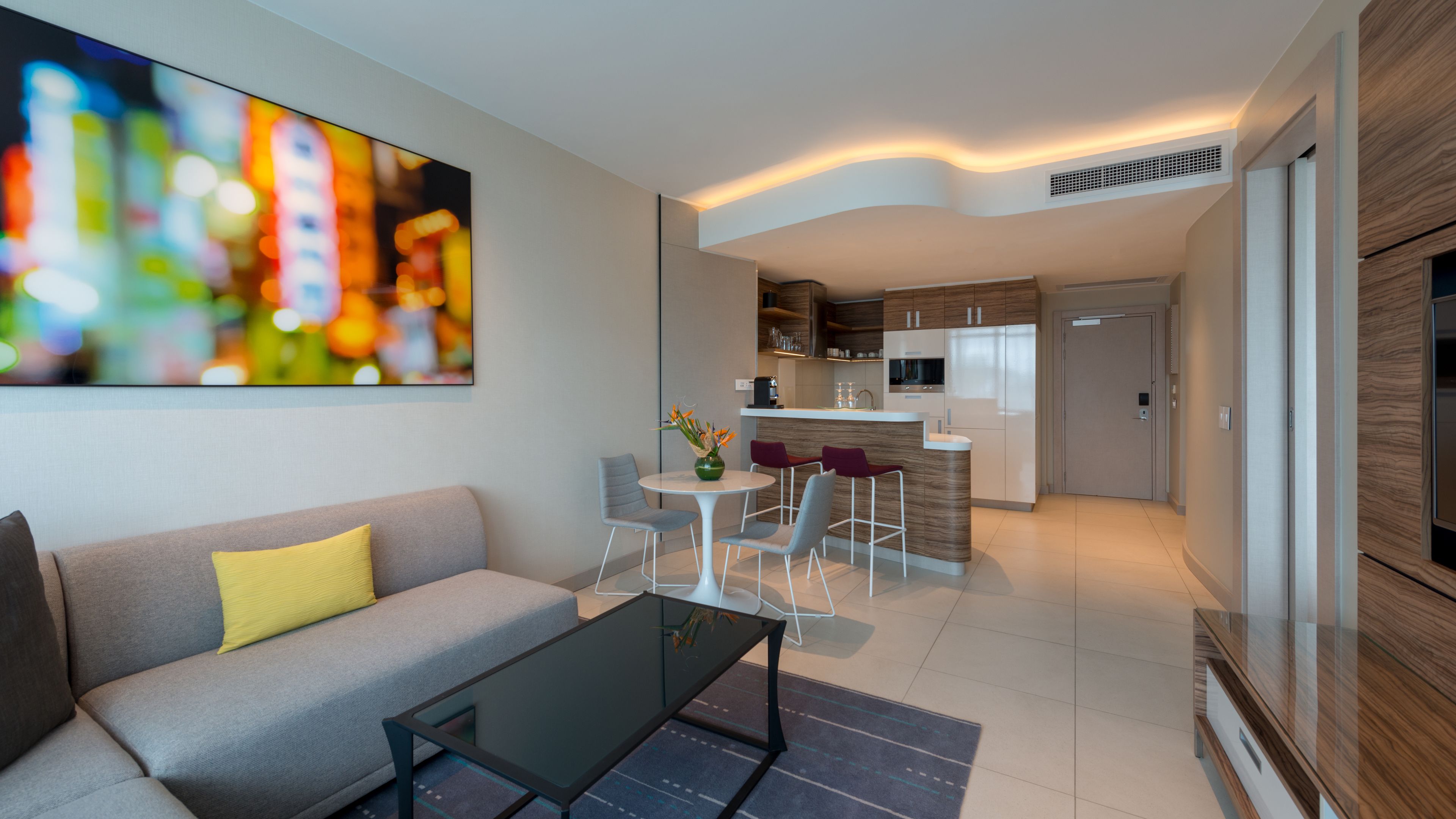 A modern living room with a sofa, a glass coffee table, and a dining area with chairs. A kitchen is visible in the background with cabinets and a countertop.
