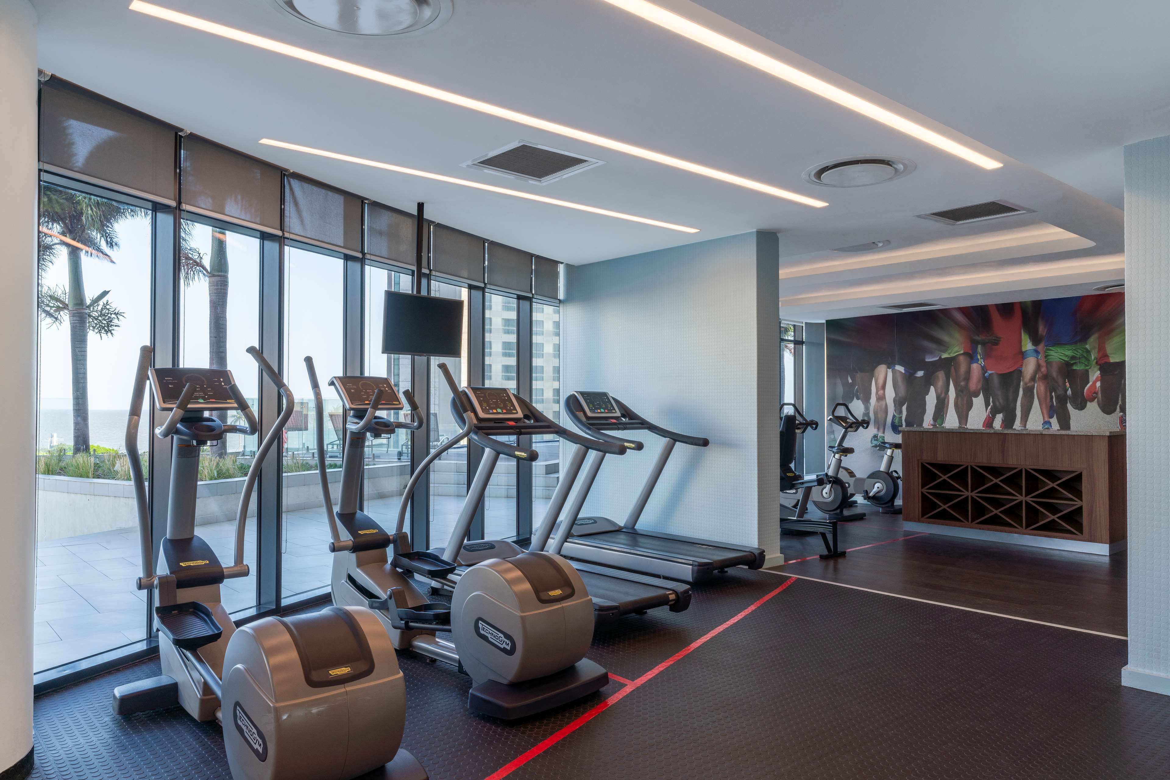 A modern gym with treadmills, ellipticals, and stationary bikes, large windows with a view, and a mural on the wall.