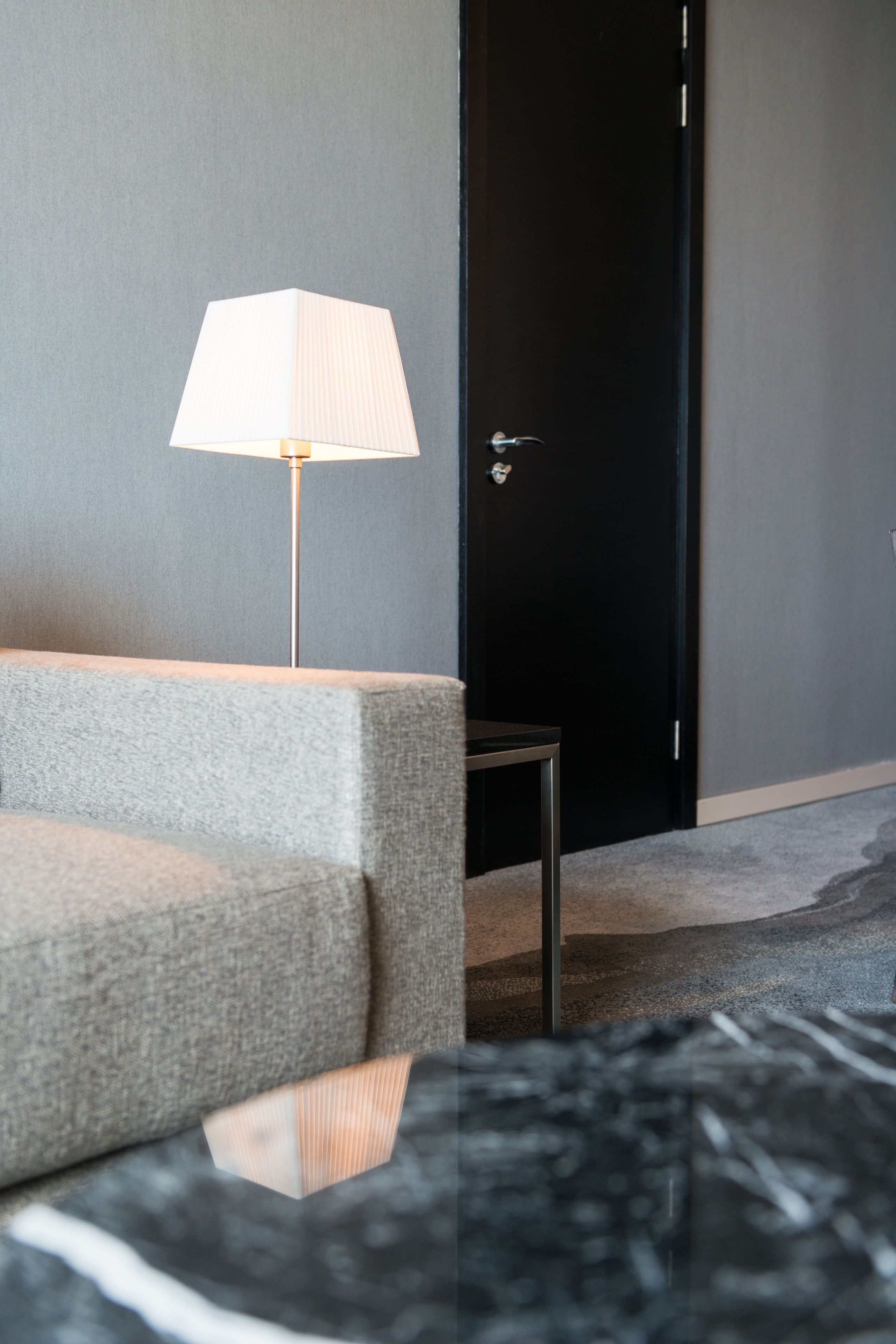 A modern hotel room with a grey sofa, a standing lamp, and a black door.
