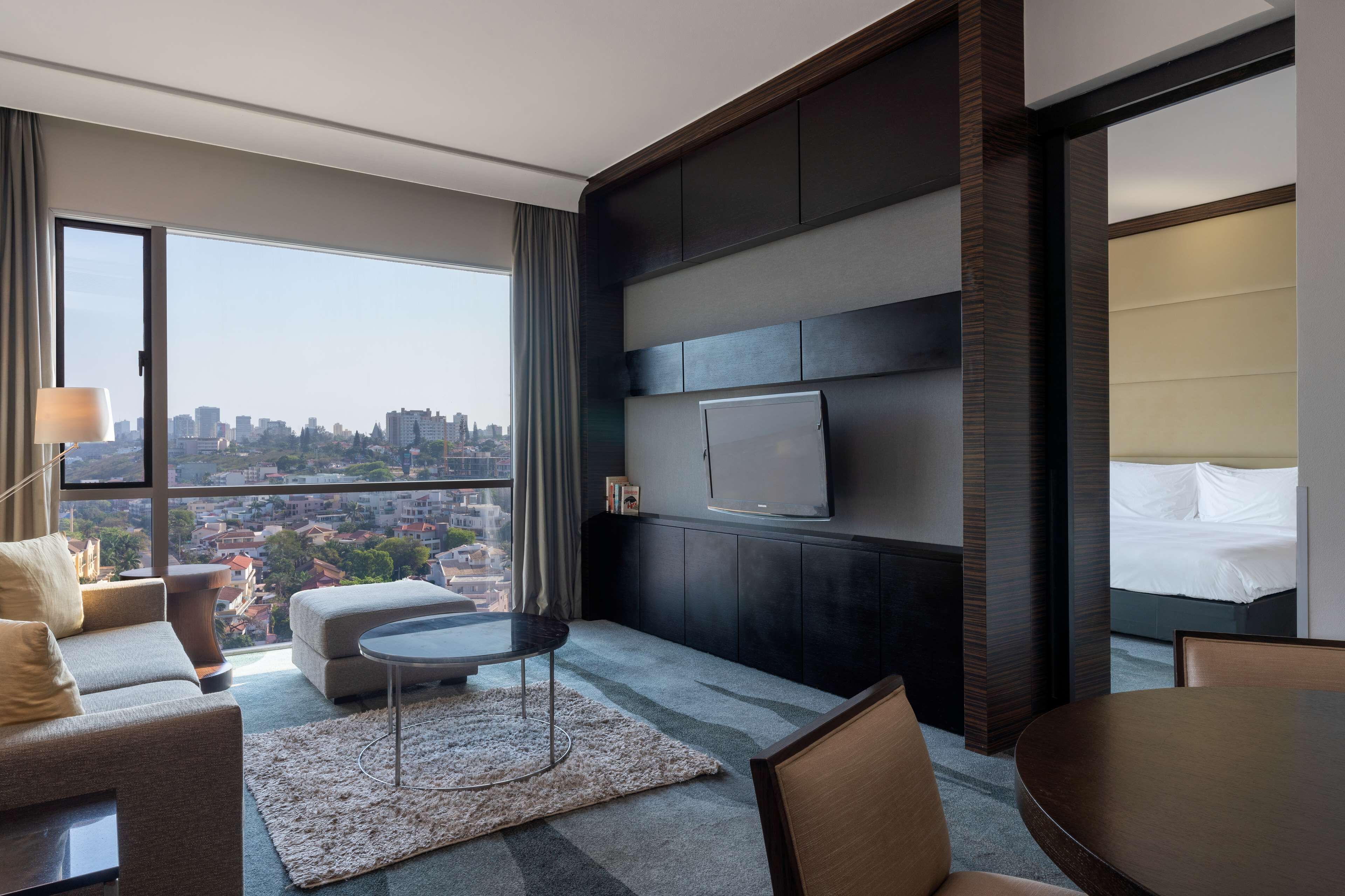 Quarto de hotel moderno com uma grande janela que oferece vista da cidade, televisão de tela plana na parede, sofá, mesa de centro redonda e cama com cabeceira.
