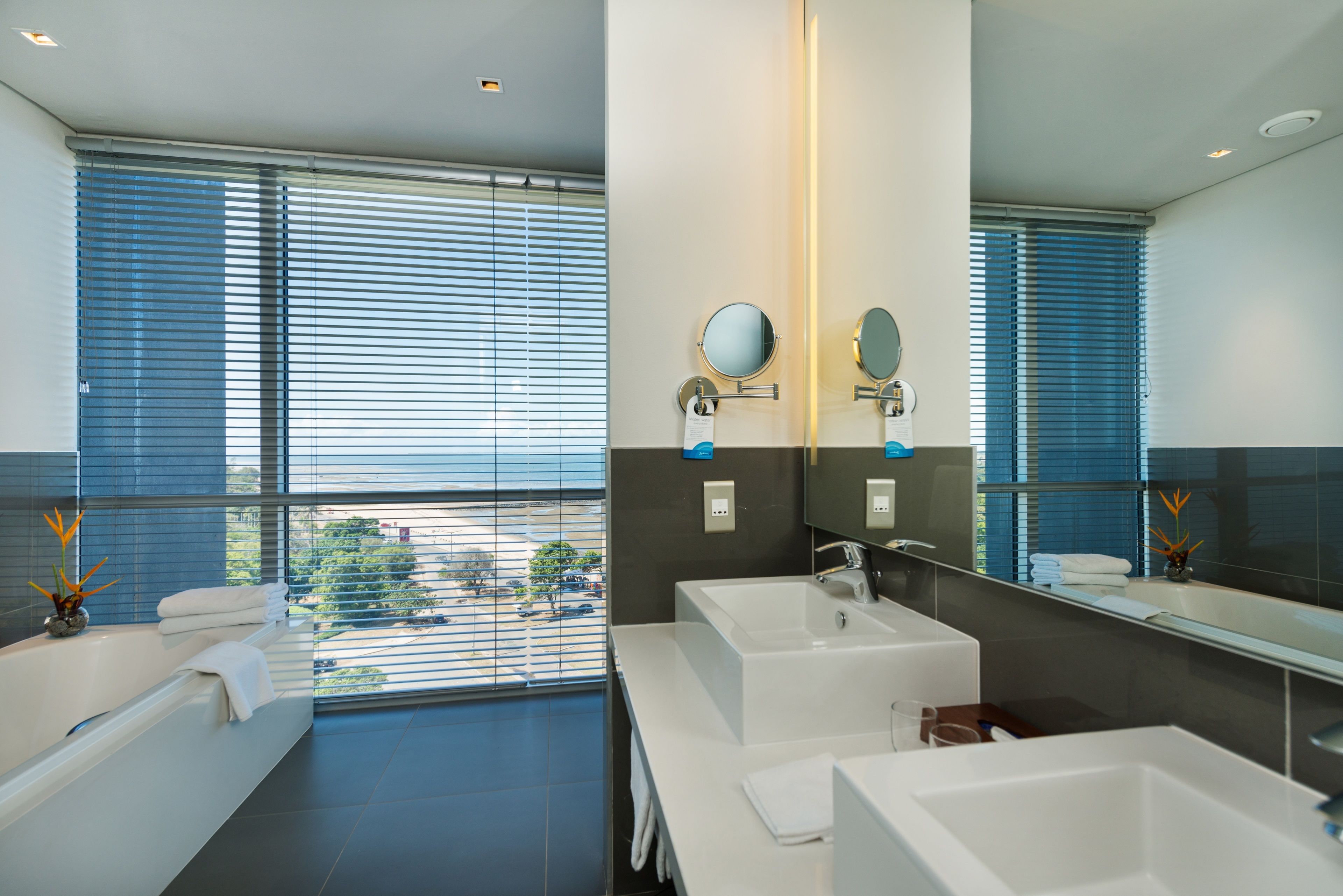 A modern bathroom with a large window, a bathtub, and two sinks.
