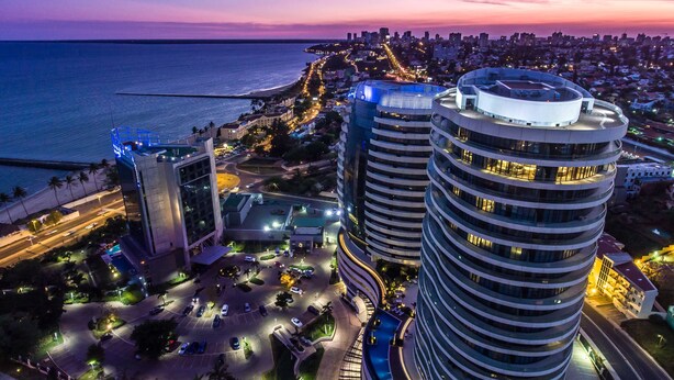 A high-rise building with a circular design, surrounded by a parking area and a coastal cityscape at dusk. - Radisson Blu Hotel & Residence, Maputo (Maputo)