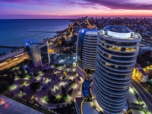 A high-rise building with a circular design, surrounded by a parking area and a coastal cityscape at dusk. - Radisson Blu Hotel & Residence, Maputo (Maputo)