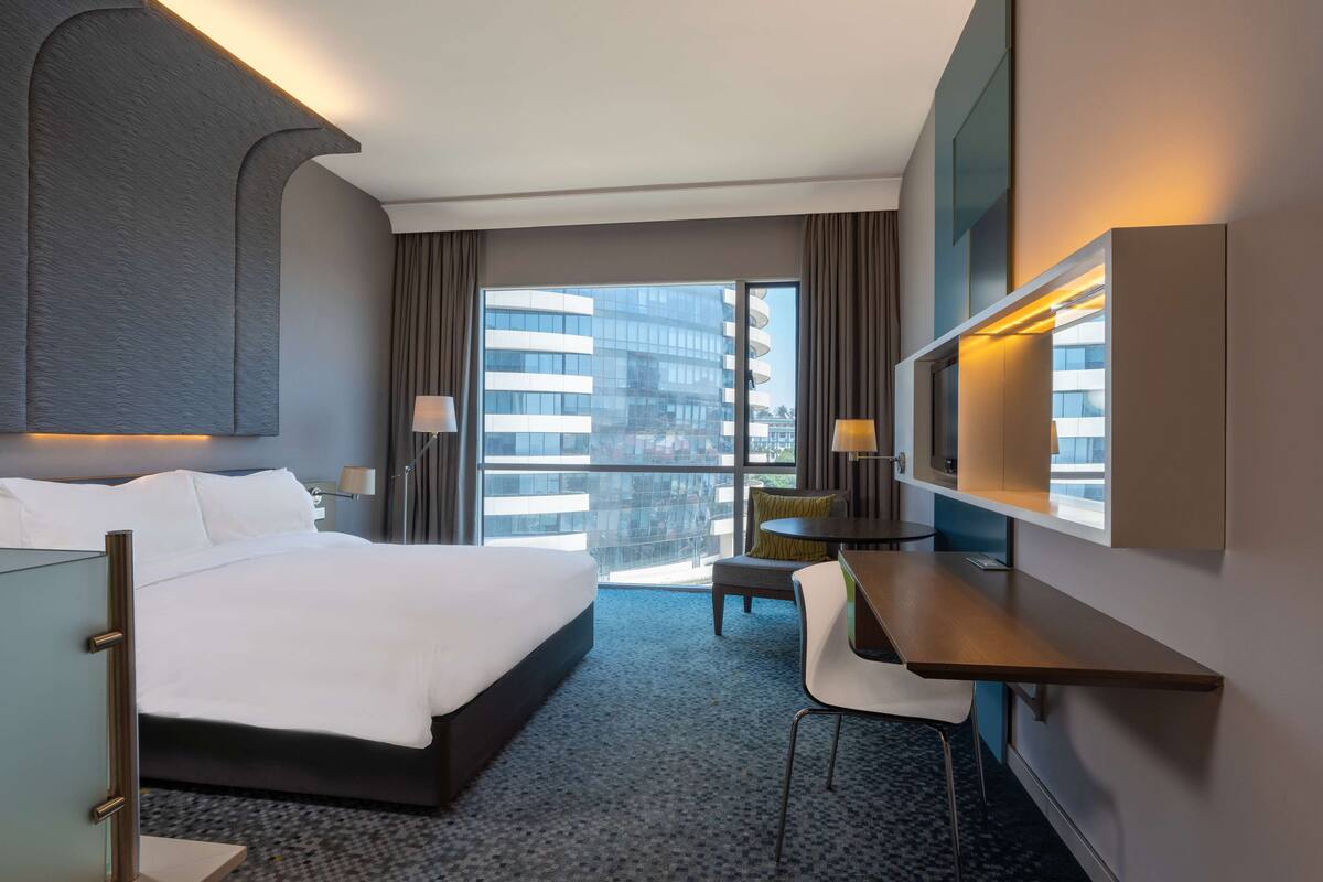 A hotel room with a large bed, a desk, a chair, a TV, and a view of a building through the window.