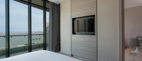 A hotel room with a large bed, a TV mounted on the wall, a balcony with a view of the sea, and a dining area with chairs.