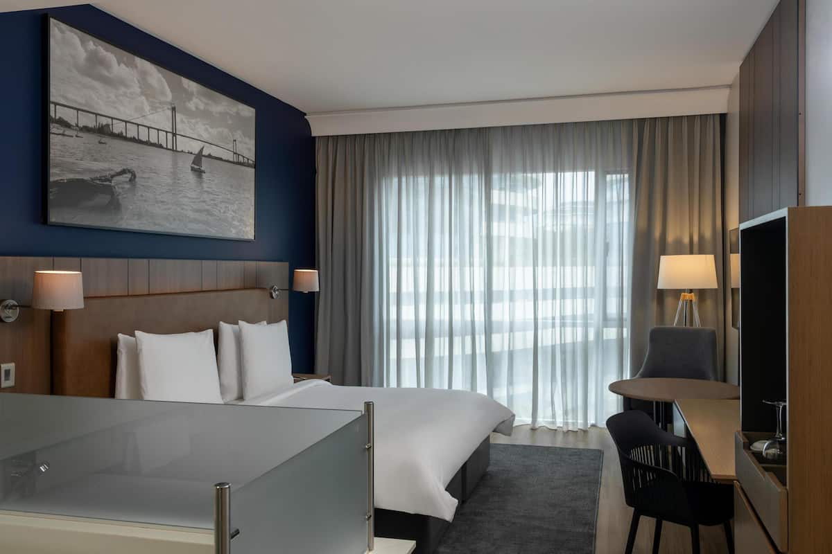 A hotel room with a bed, bedside tables, a desk with a chair, a large window with curtains, and a painting on the wall.