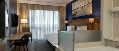 Habitación de hotel con cama, escritorio y silla, televisor, un cuadro en la pared, ventana con cortinas y bañera.